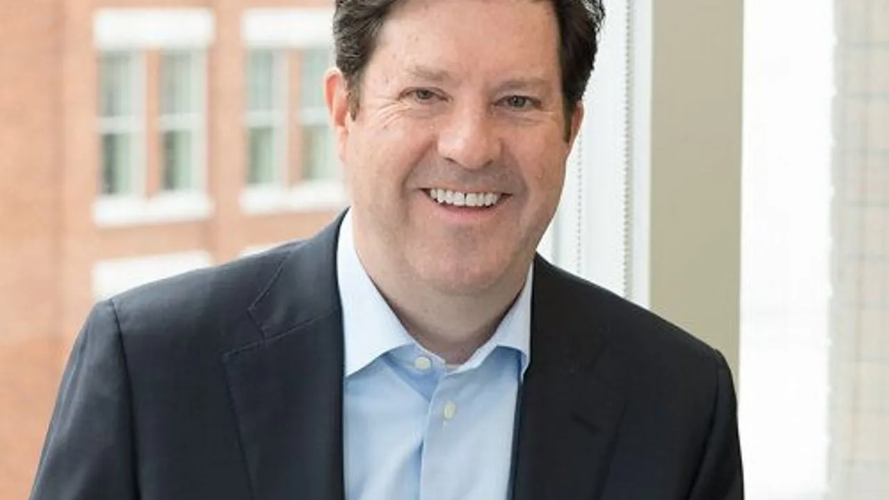 A smiling person in a dark suit and light blue shirt stands by a window.