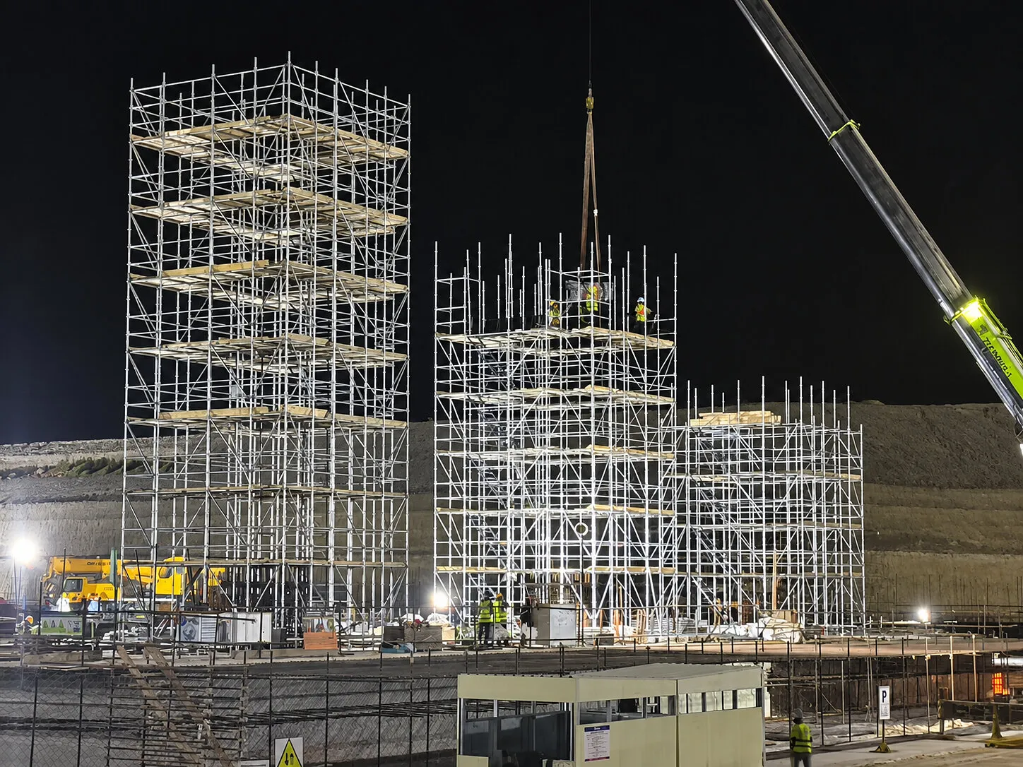 Scaffolding towers and a crane at a floodlit construction site at night.