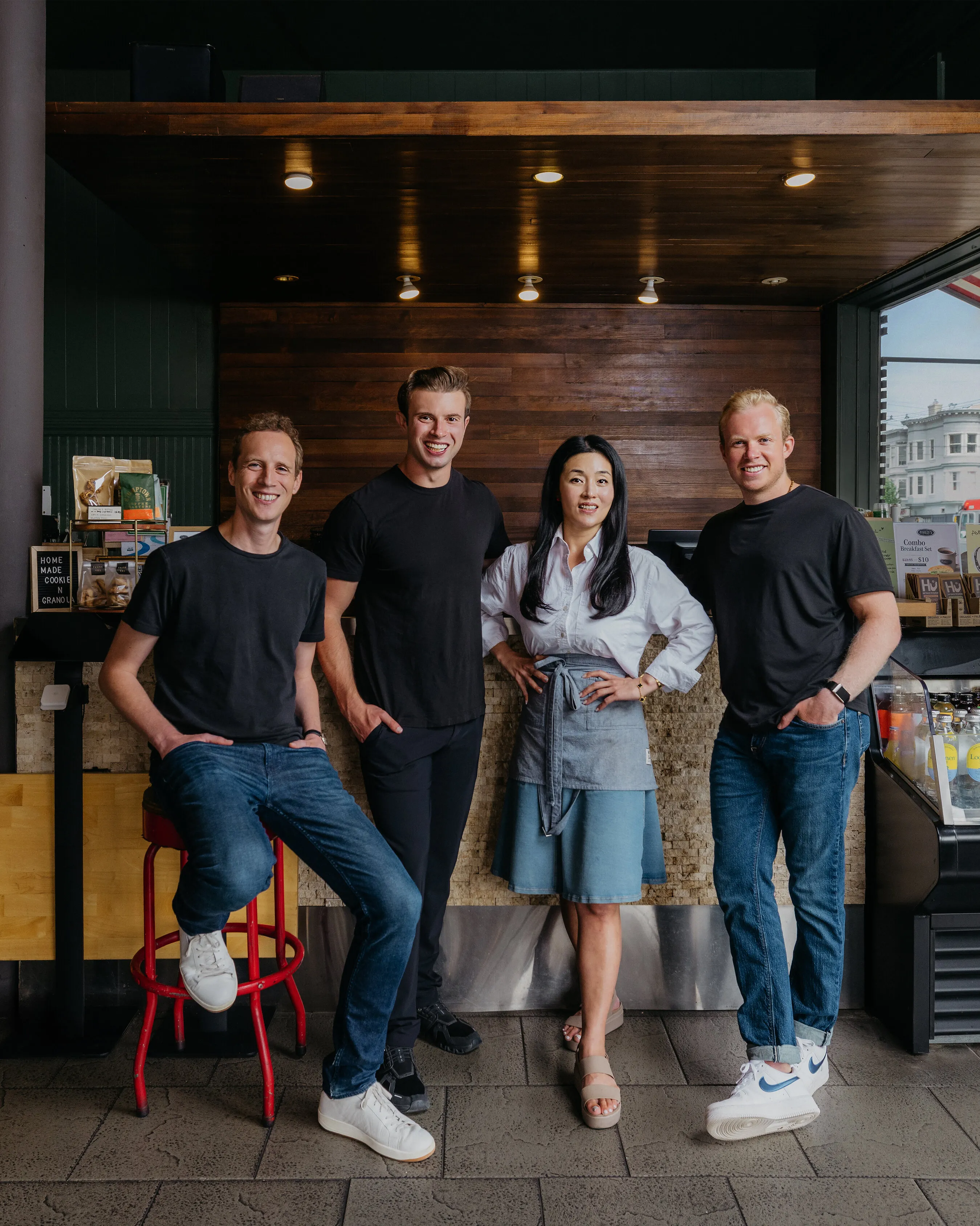 Four people in a café, one seated on a red stool and three standing.