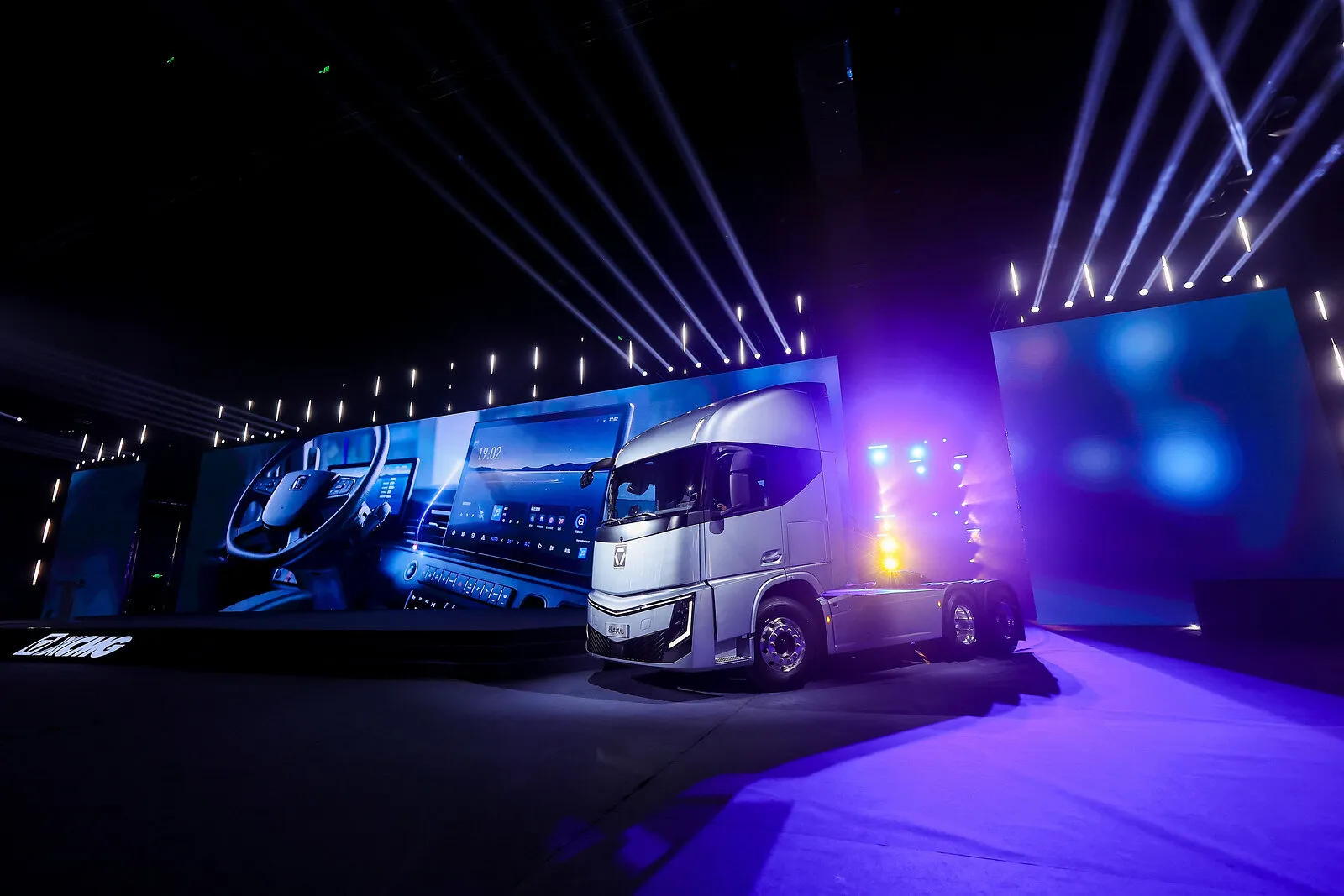 Silver semi-truck tractor displayed on a stage with large screens and purple-blue lights.