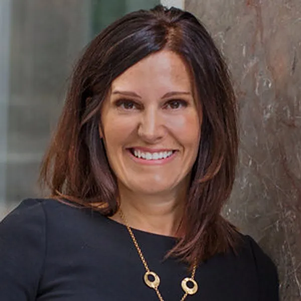 A smiling person with dark brown hair wears a black top and gold necklace beside a stone wall.