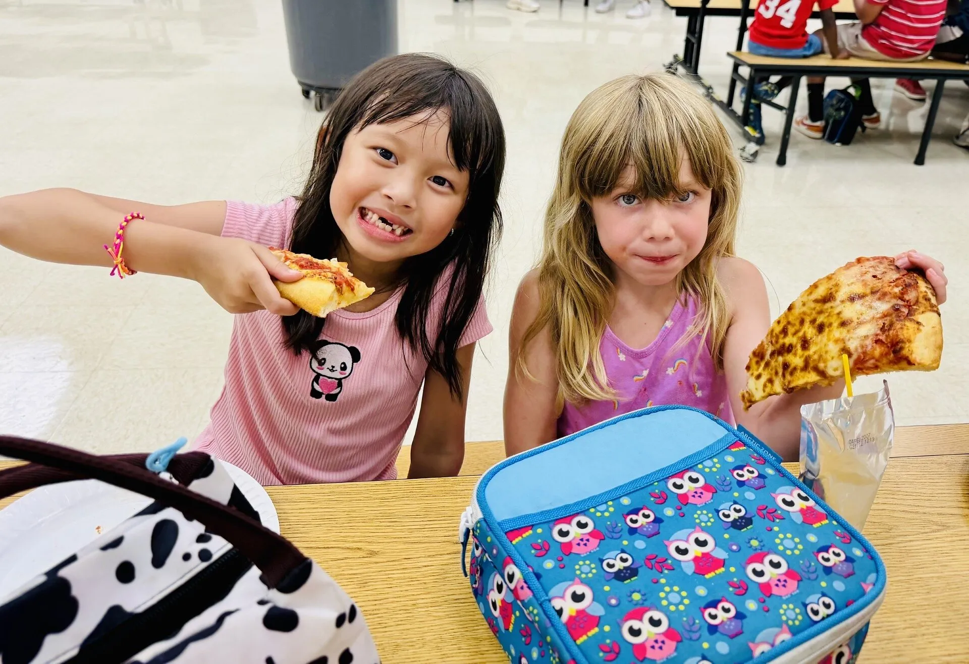 Two girls hold slices of pizza over a cafeteria table with colorful lunch bags.