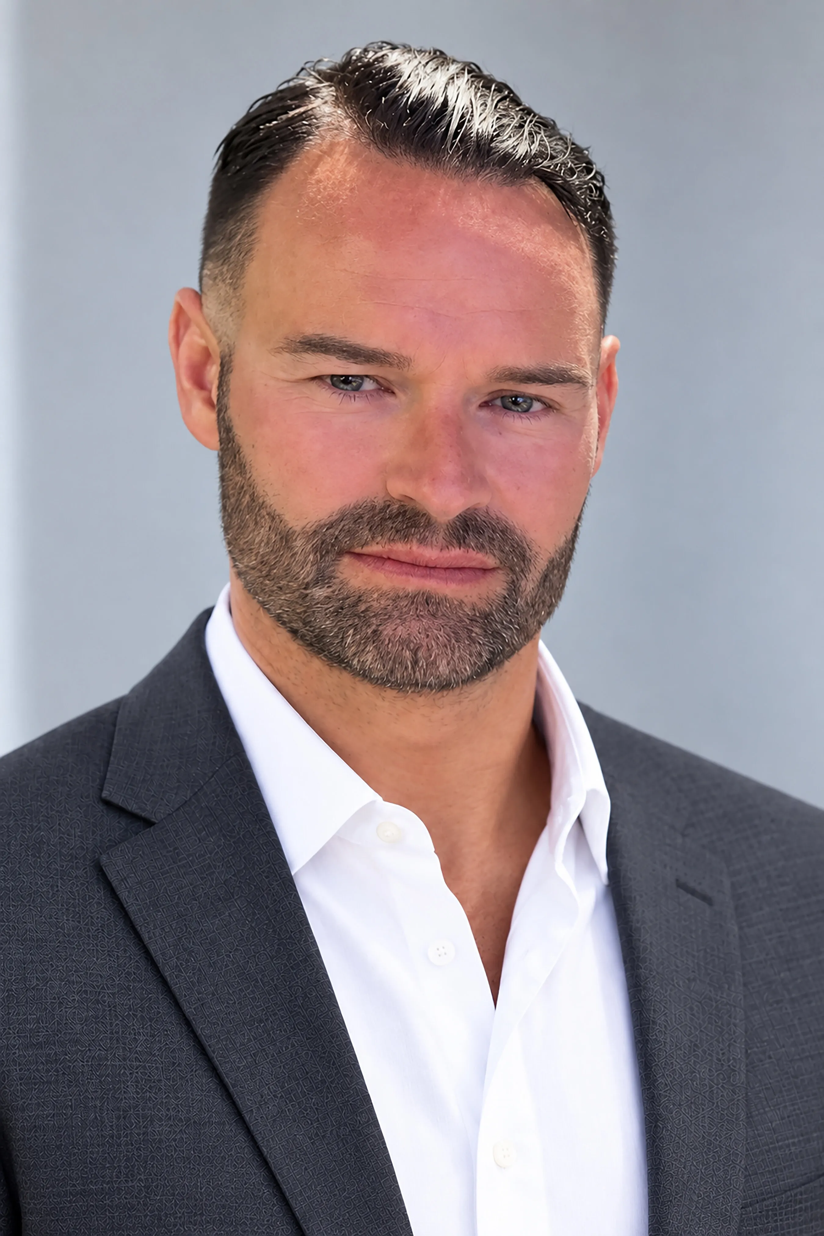 A bearded man in a dark suit jacket and white shirt against a gray background.