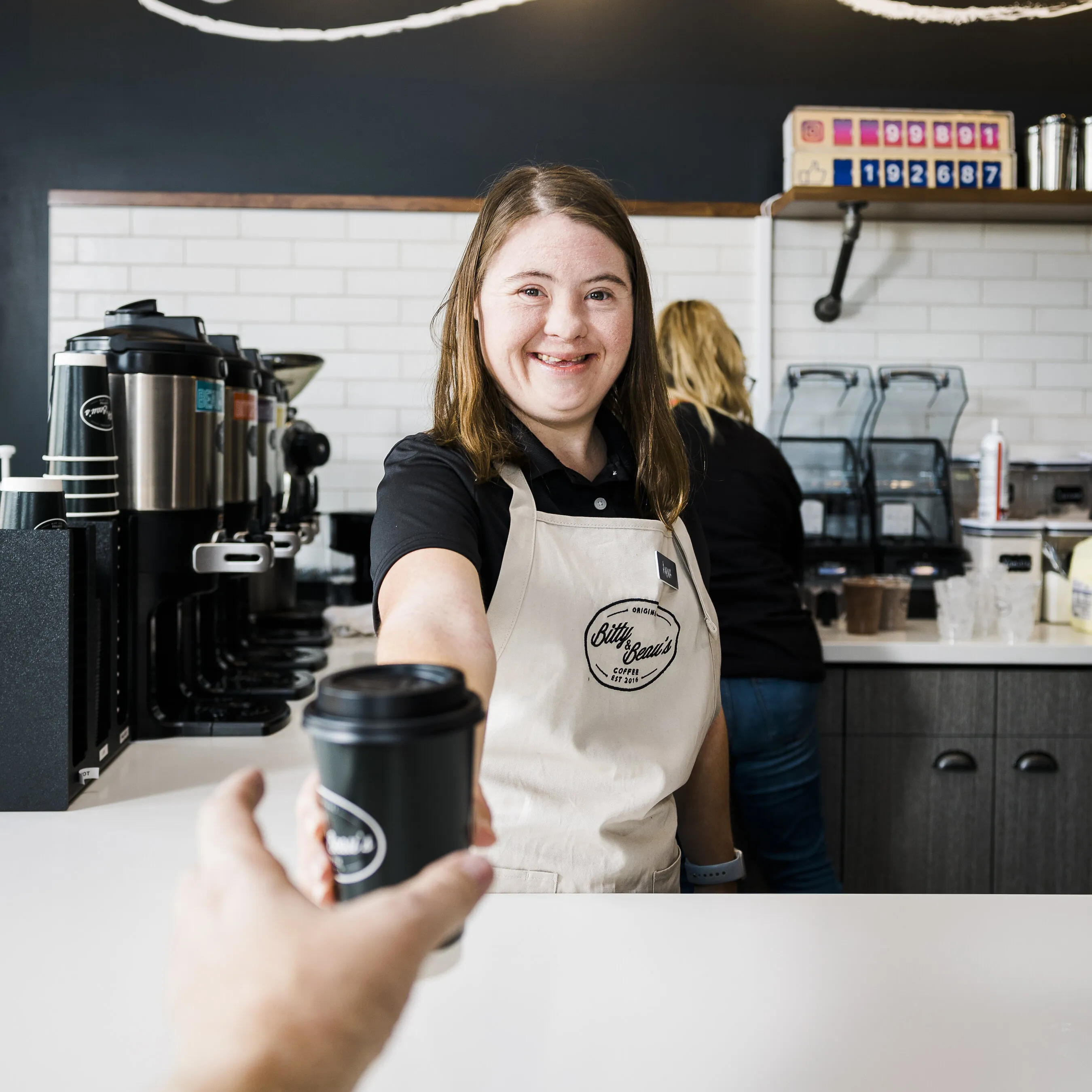 Barista in a Bitty & Beau’s Coffee apron hands a customer a takeaway coffee cup.