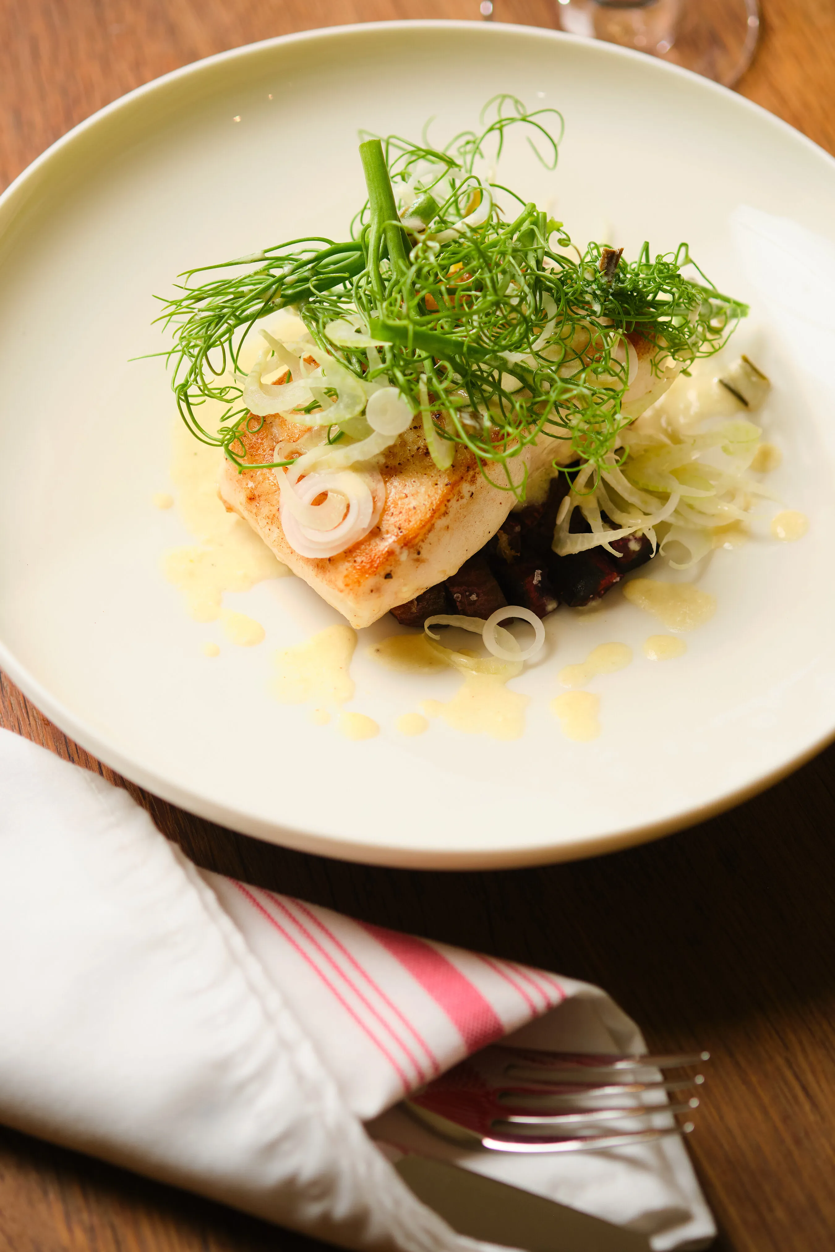 Seared white fish with greens, sliced shallots and creamy sauce on a white plate.