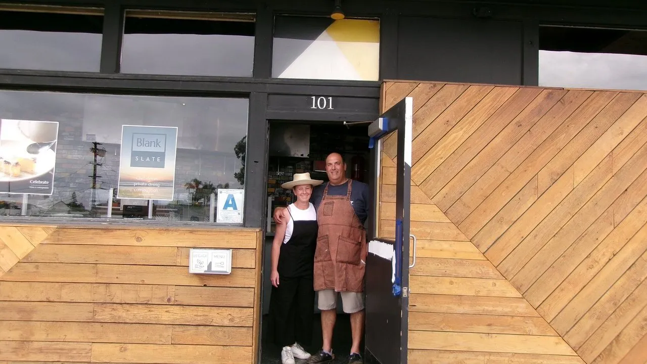 Two people in aprons stand at the entrance of a wood-paneled restaurant.