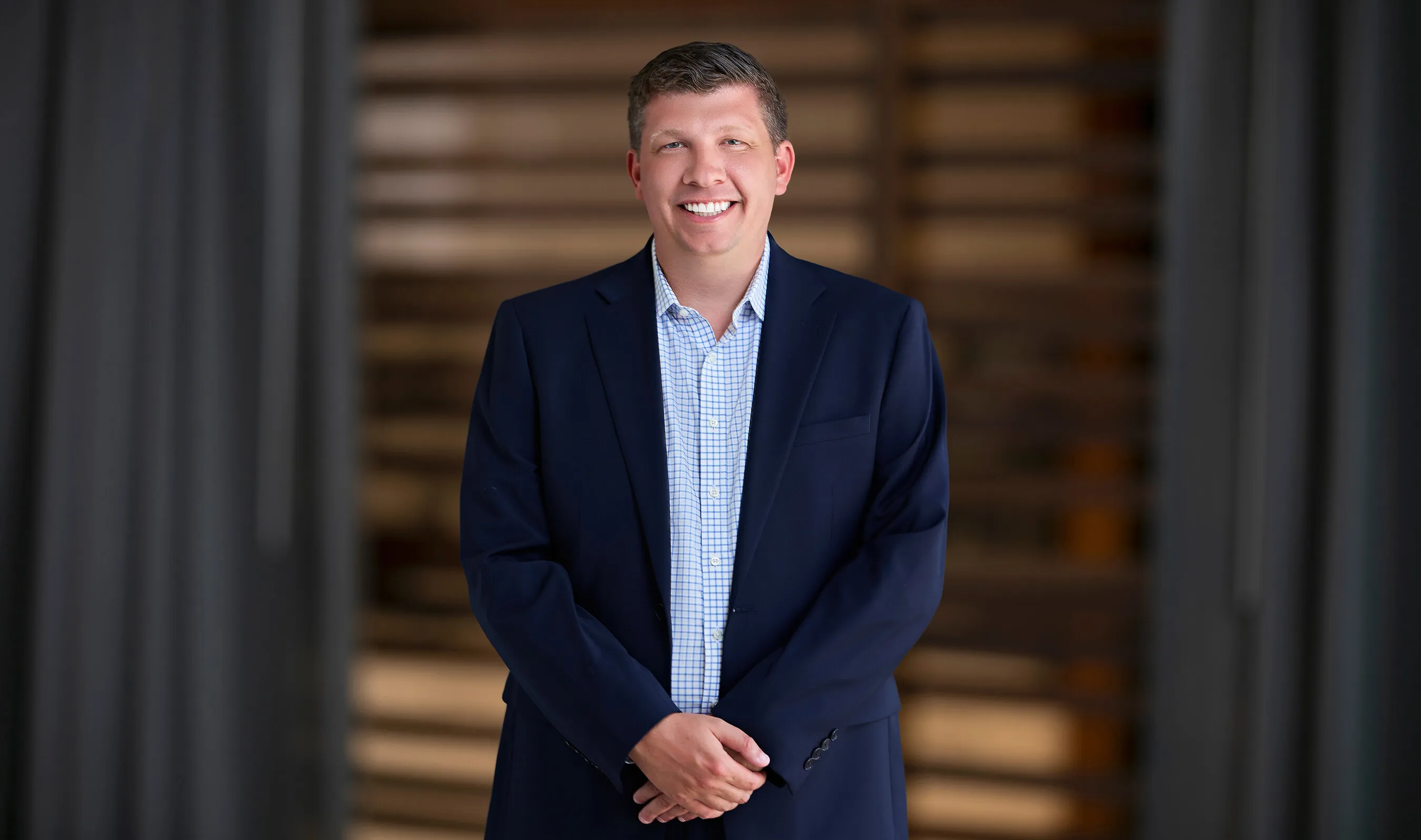Cory Boggs smiling in a navy blazer and checked shirt in front of a blurred wooden background.