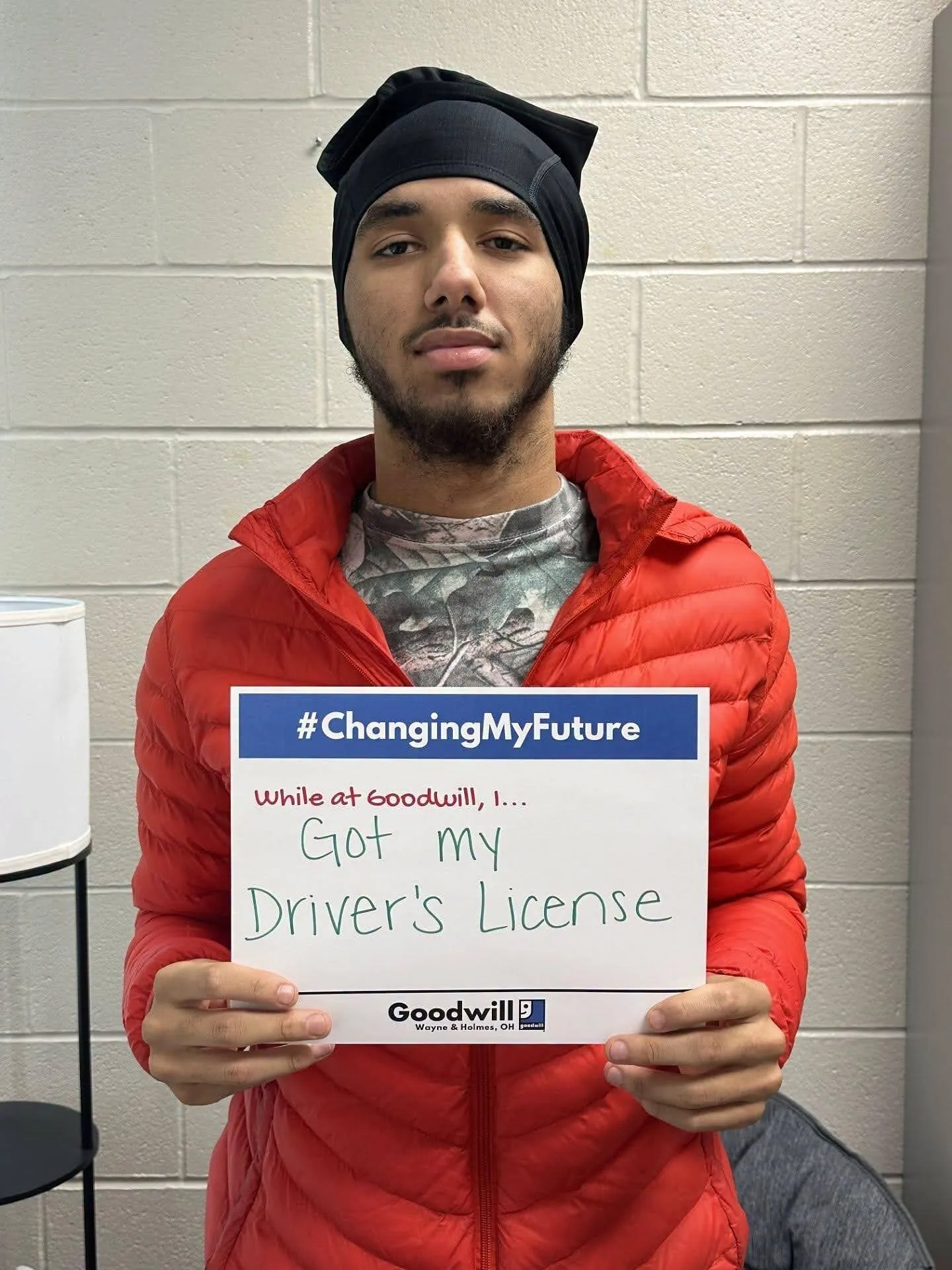 A man in a red jacket holds a Goodwill sign reading “Got my Driver’s License.”