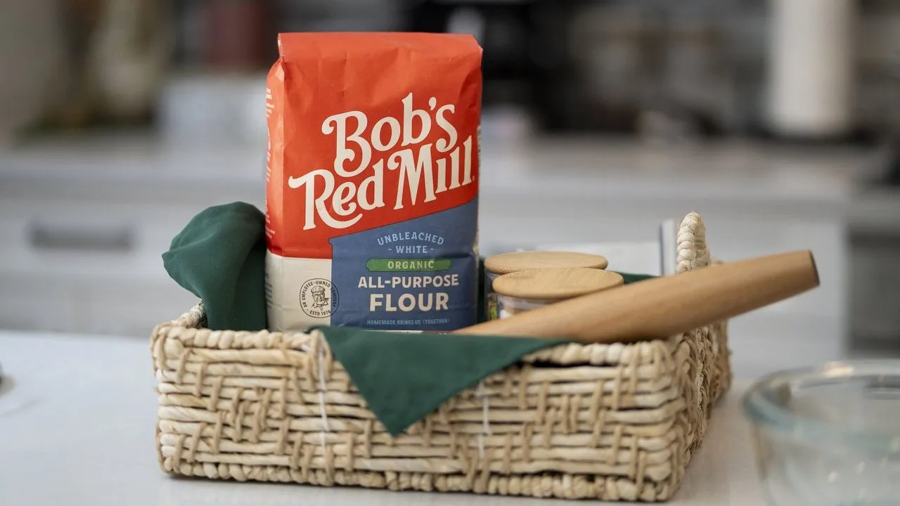 A bag of flour, rolling pin and jars in a woven basket on a kitchen counter