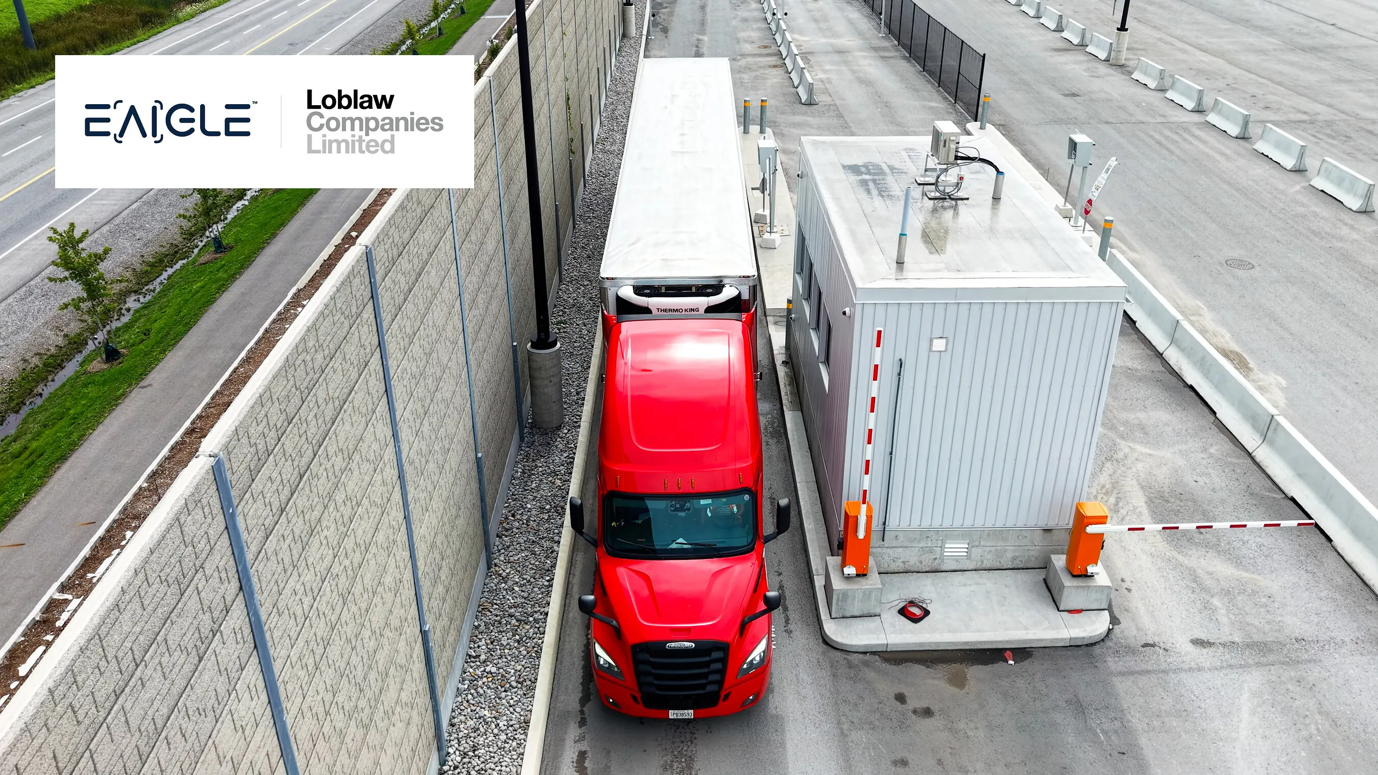 Red semi-truck passing a security checkpoint beside a gray inspection building and barrier gate.