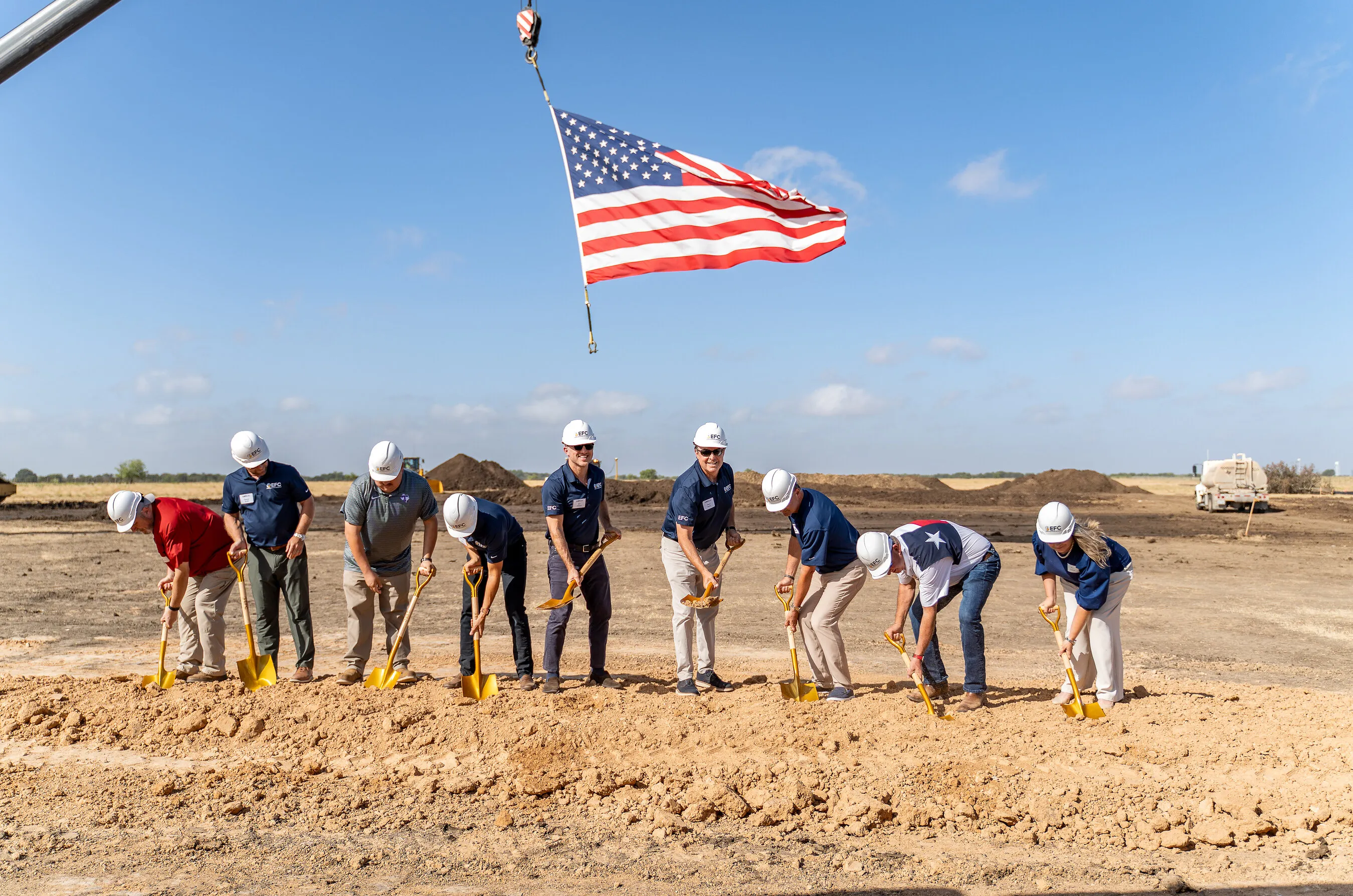 Nine people in hard hats break ground with gold shovels beneath an American flag.
