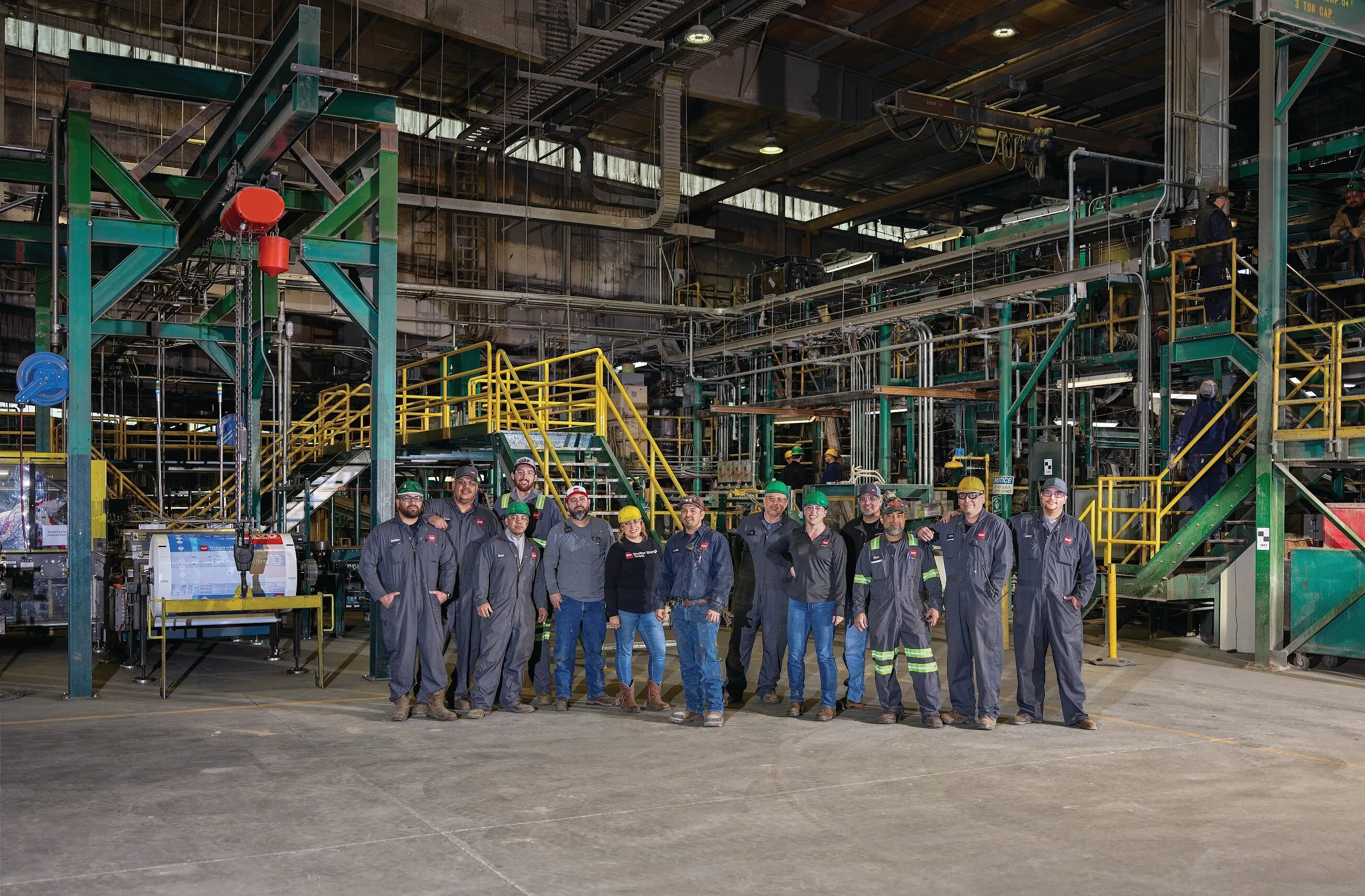 A group of GAF workers, some in hard hats and coveralls, stands inside an industrial manufacturing facility.