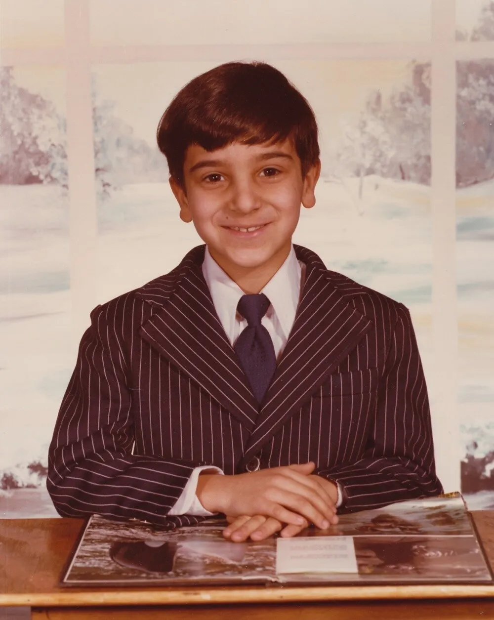 John Katsimatides as a boy in a pinstripe suit and tie, seated at a table with an open book.