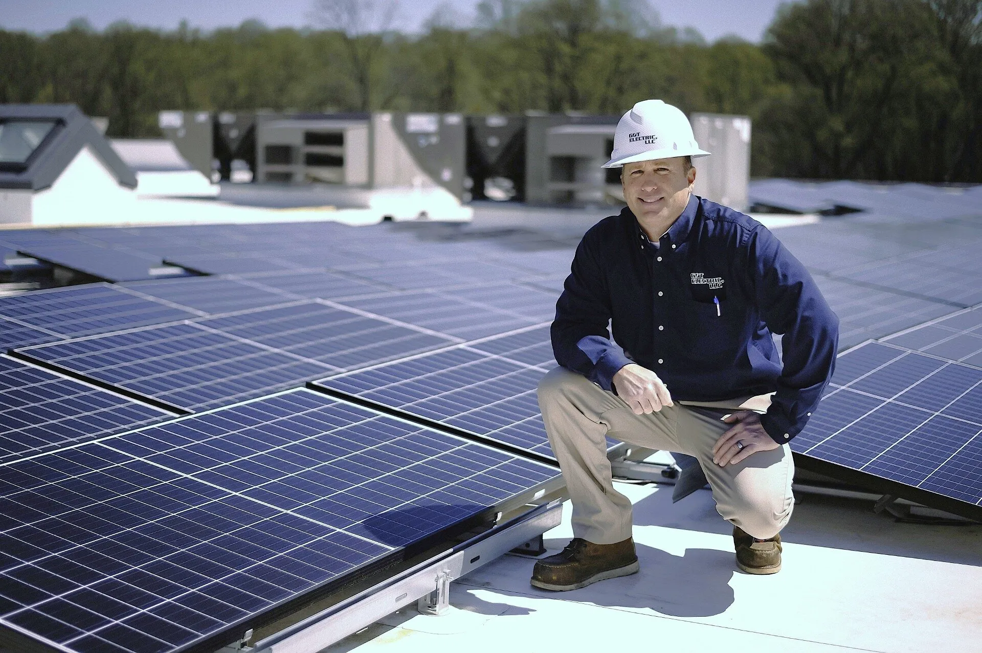 A worker in a hard hat kneels beside rooftop solar panels.