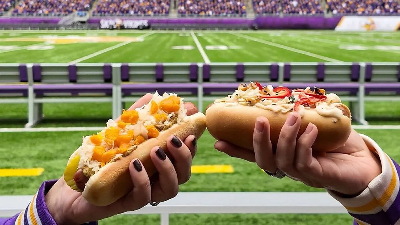 Two hands hold loaded hot dogs in front of a football field and stadium seating.