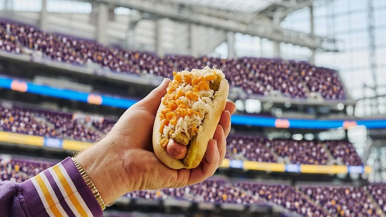 A hand holds a SPAM Dog topped with sauerkraut and orange chips in a stadium.