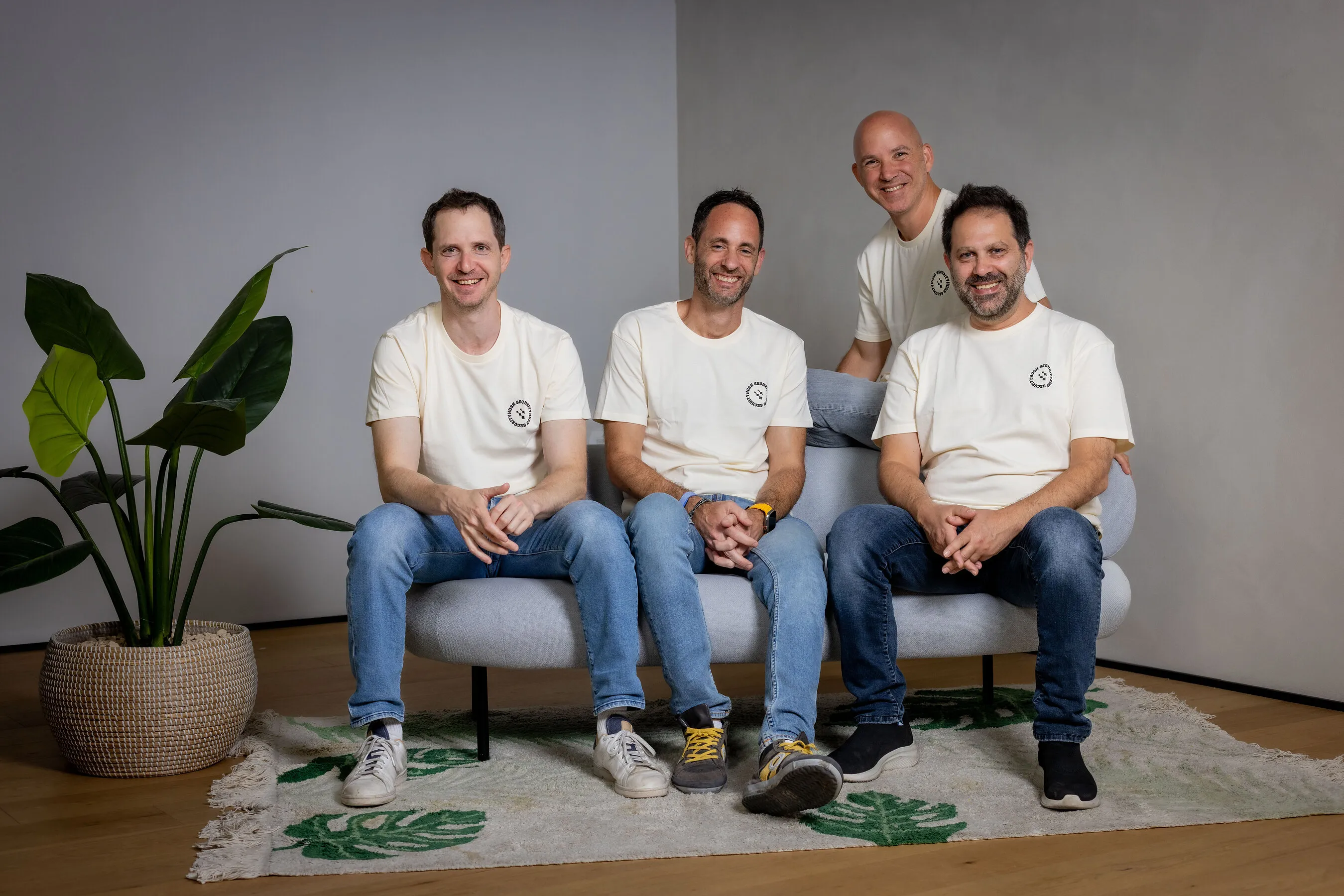 Four men in white T-shirts and jeans sit and stand around a sofa beside a potted plant.