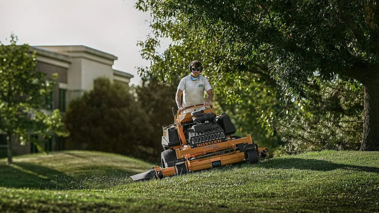 A worker operates a Hustler lawn mower on a grassy slope beneath trees.