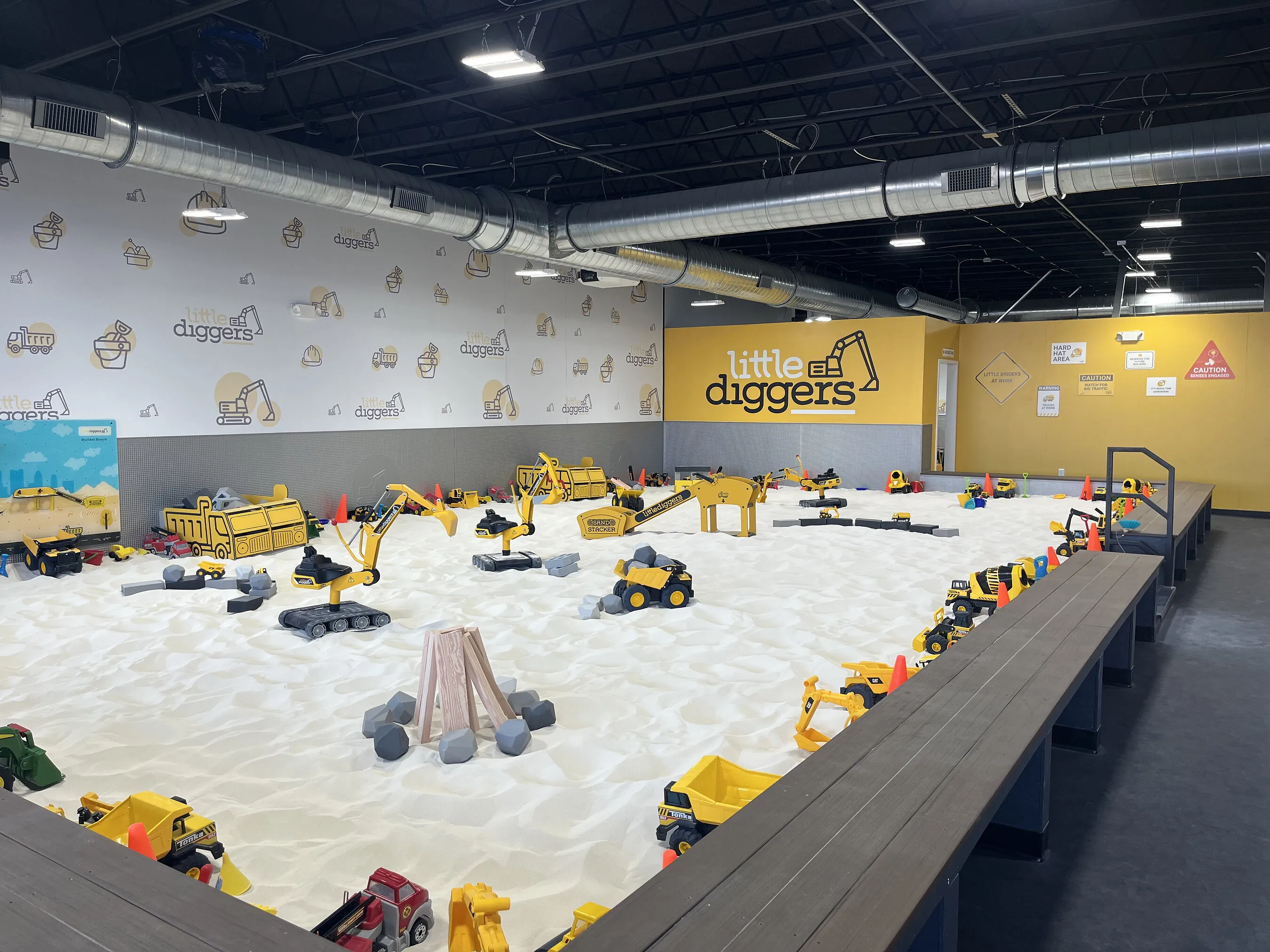 Indoor sand play area filled with toy construction vehicles beneath a Little Diggers sign