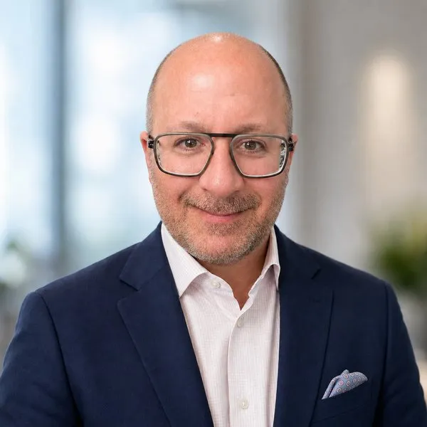 A bald man wearing glasses, a navy suit jacket and a white shirt smiles at the camera.