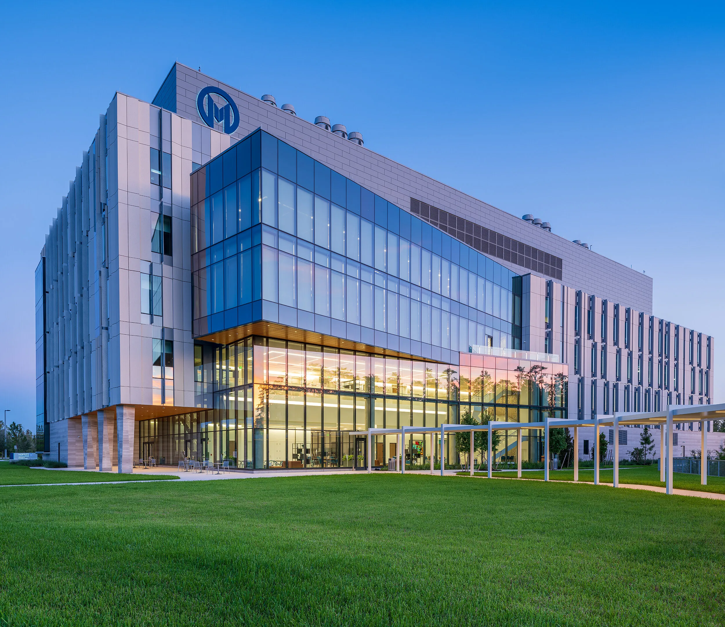 Modern glass-fronted building with illuminated interior beside a lawn at dusk