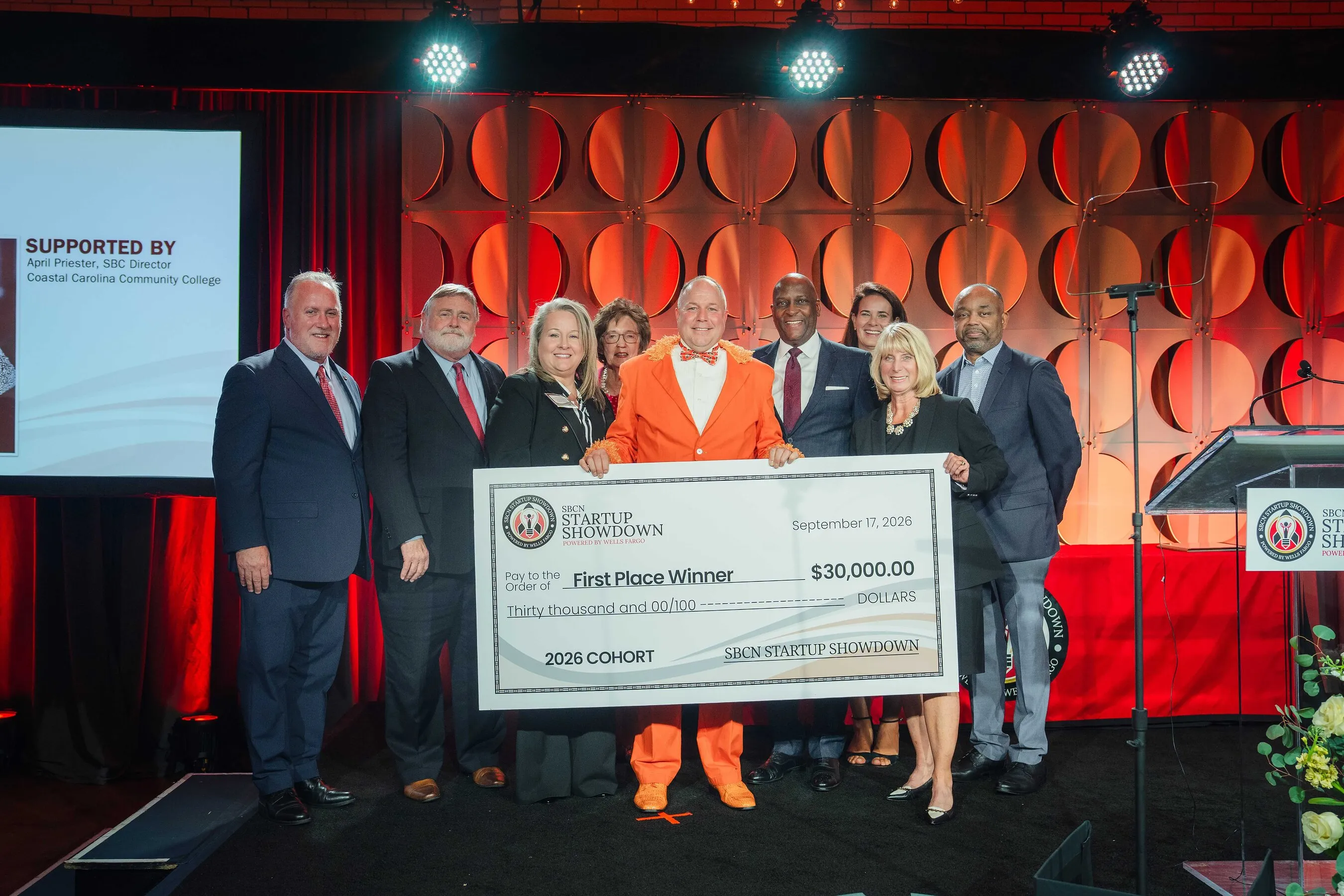 Nine people stand onstage behind a $30,000 Startup Showdown first-place winner check, with winner Dan Spangler in an orange suit in the middle.