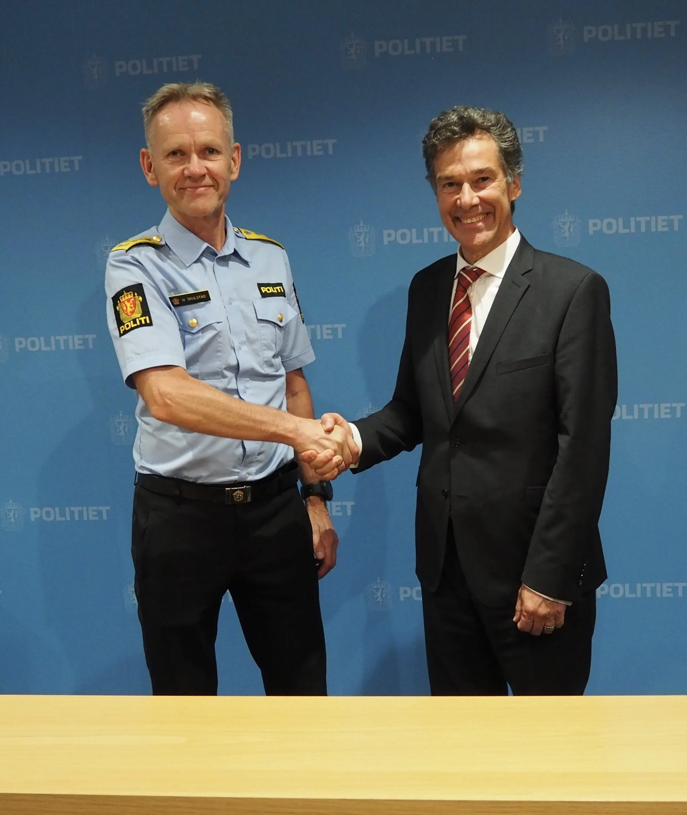 A police officer and a man in a suit shake hands in front of a Politiet backdrop.