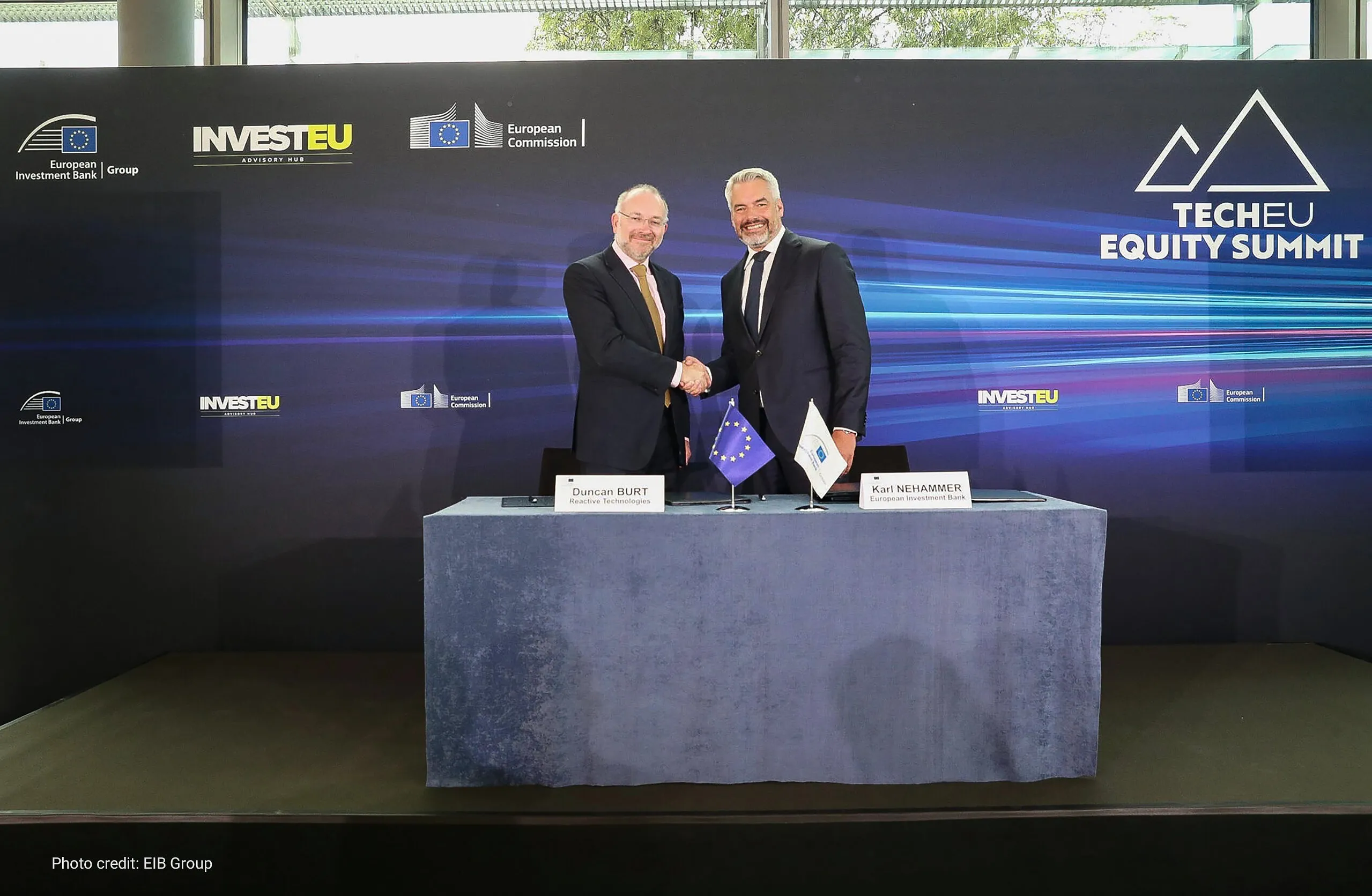 Two men shake hands behind a table with an EU flag and an EIB flag at the TechEU Equity Summit.