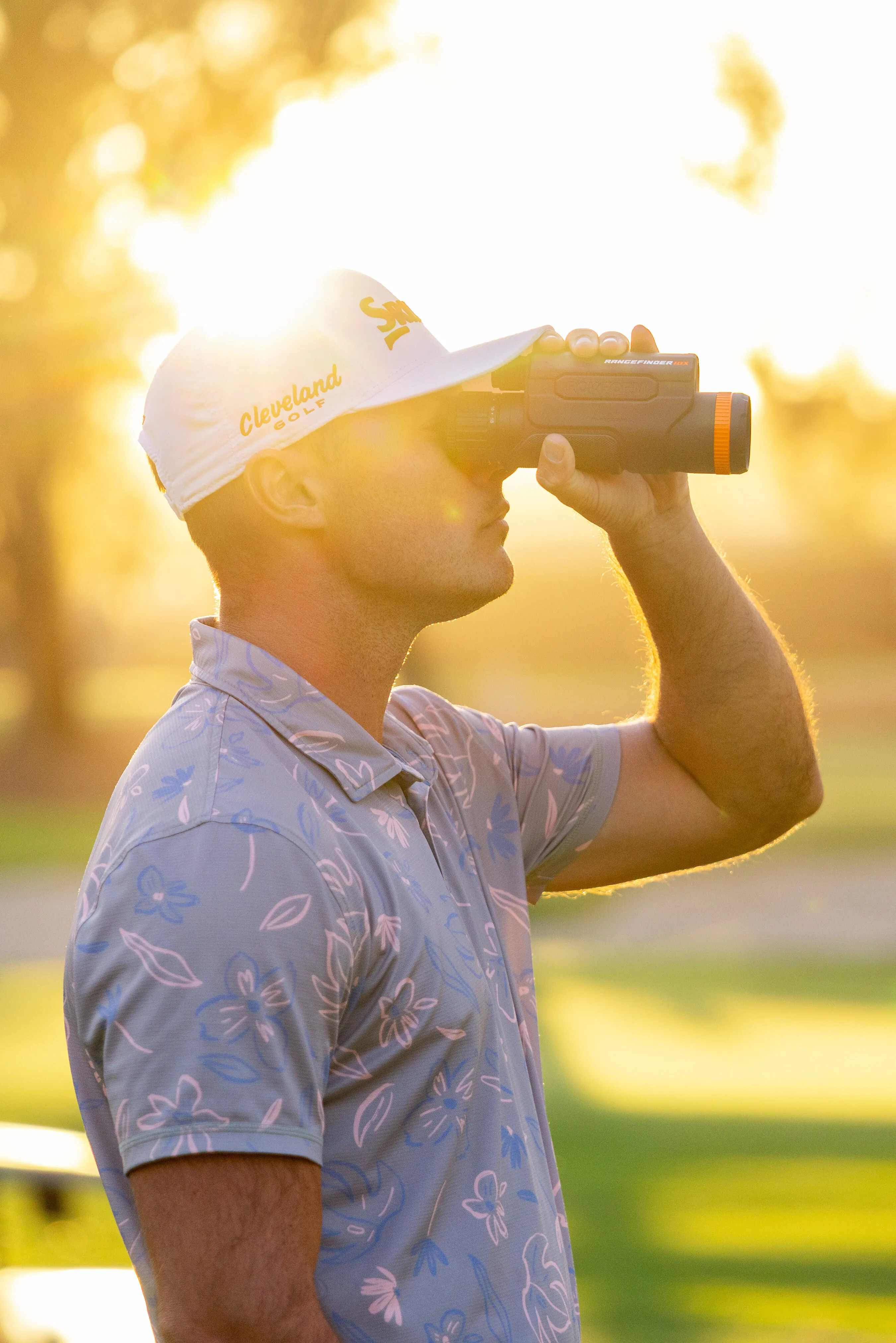 A golfer uses a rangefinder while standing on a sunlit course.