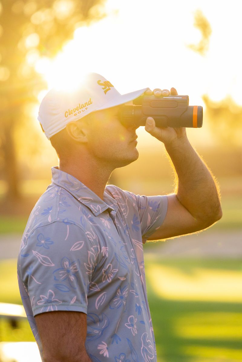 A golfer uses a rangefinder while standing on a sunlit course.