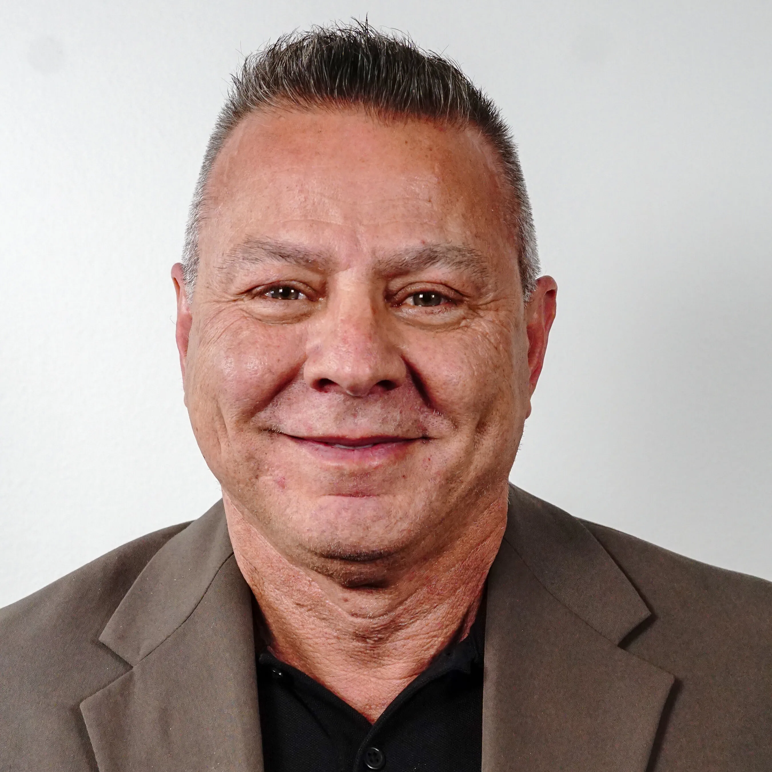 A close-up headshot of a smiling man with short grey hair in a brown jacket and black shirt.