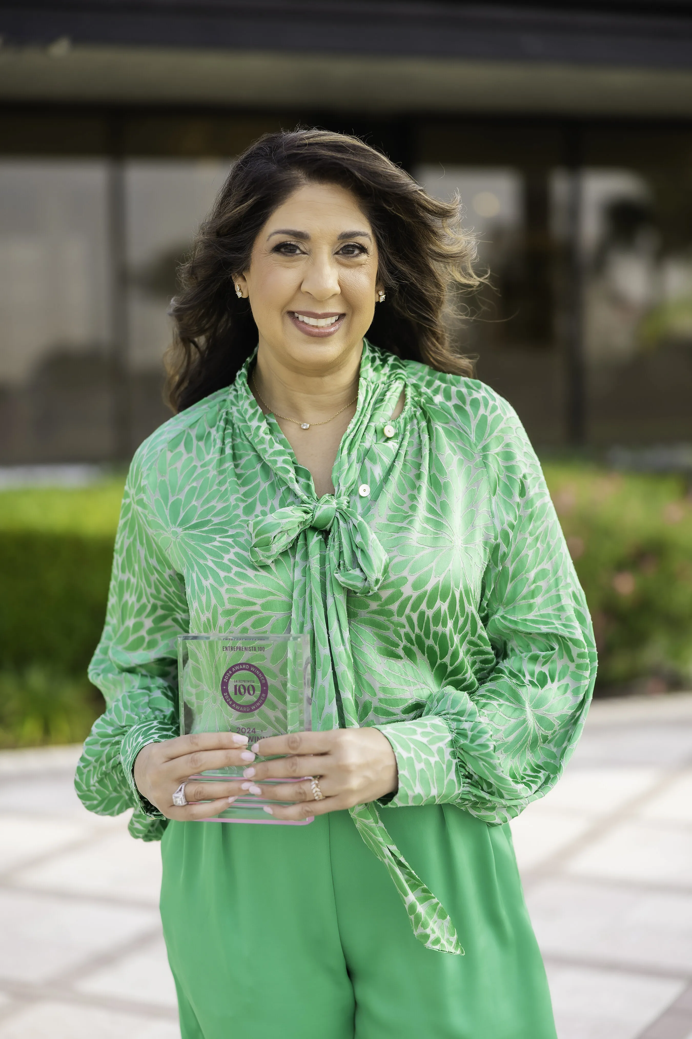 Ruchi Pinniger holds an award plaque outdoors, wearing a green patterned blouse.