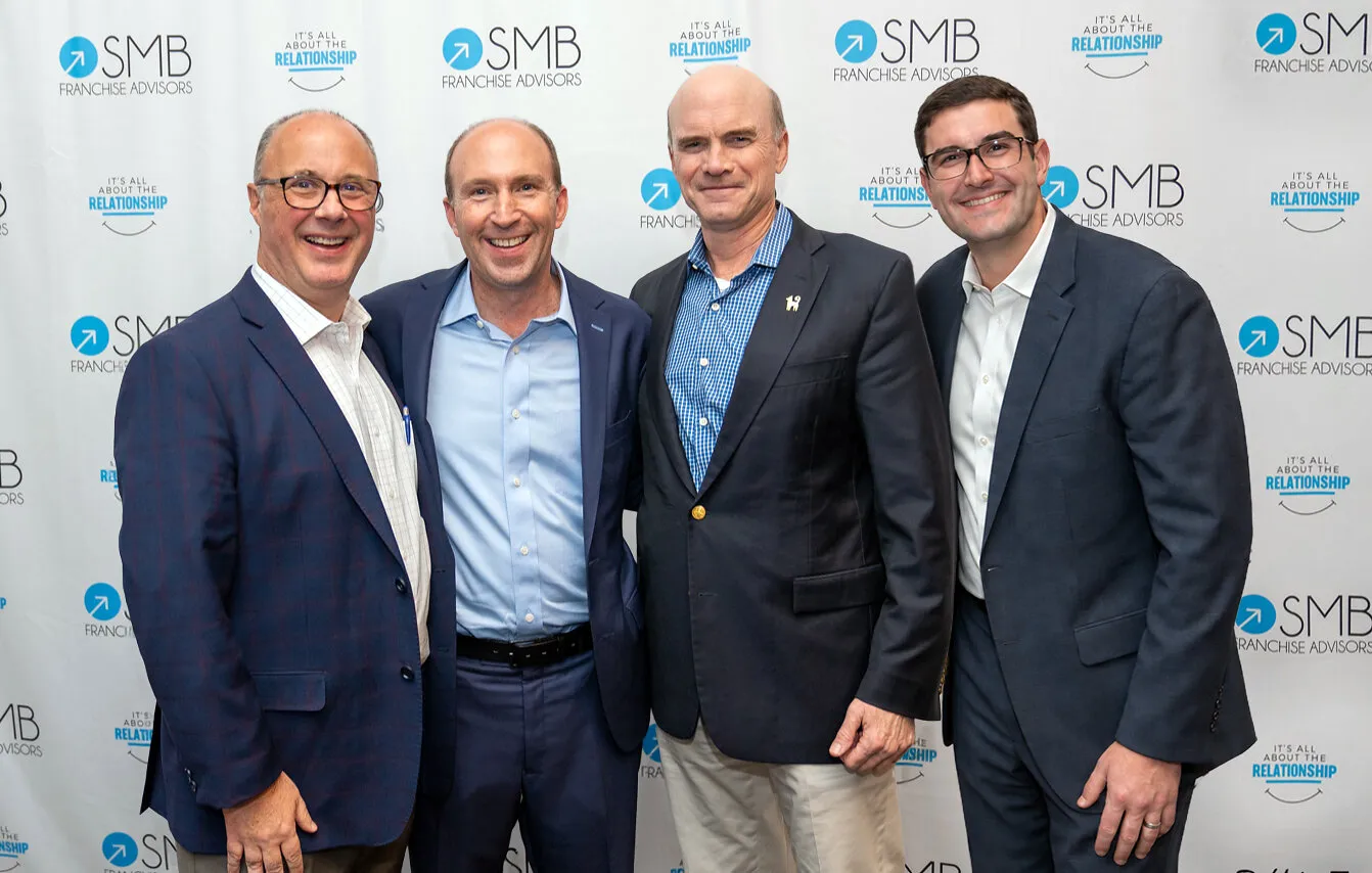 Four men in business suits pose before an SMB Franchise Advisors backdrop.