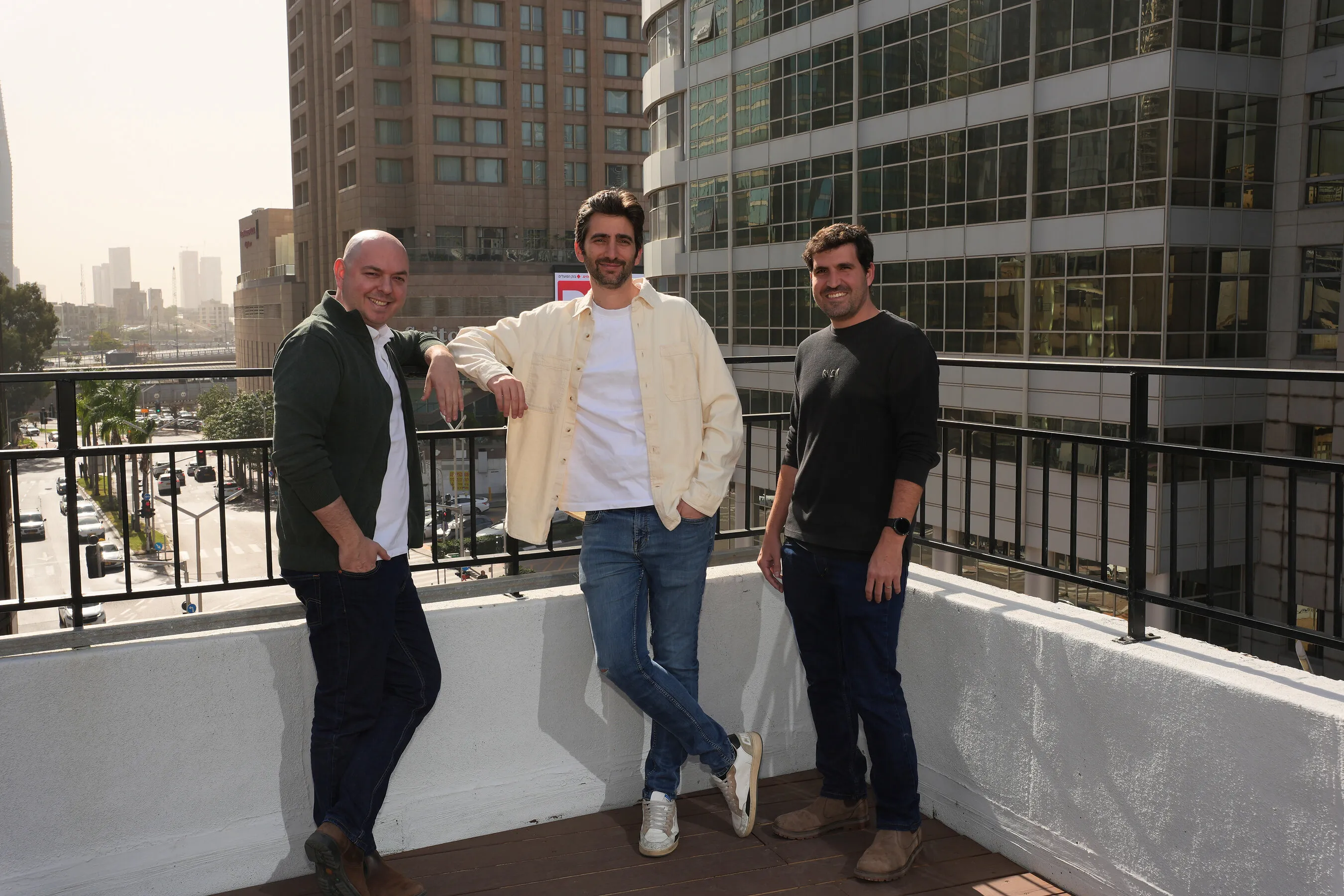 Three men stand on a rooftop terrace overlooking city streets and office buildings.