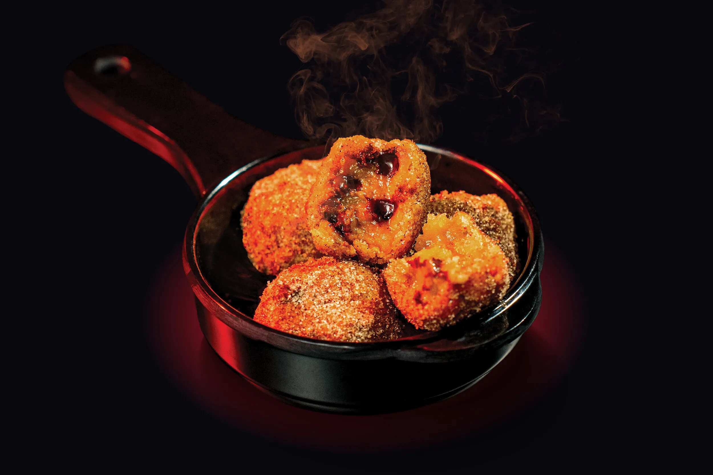 Steaming fried croquettes in a small black skillet against a dark background.