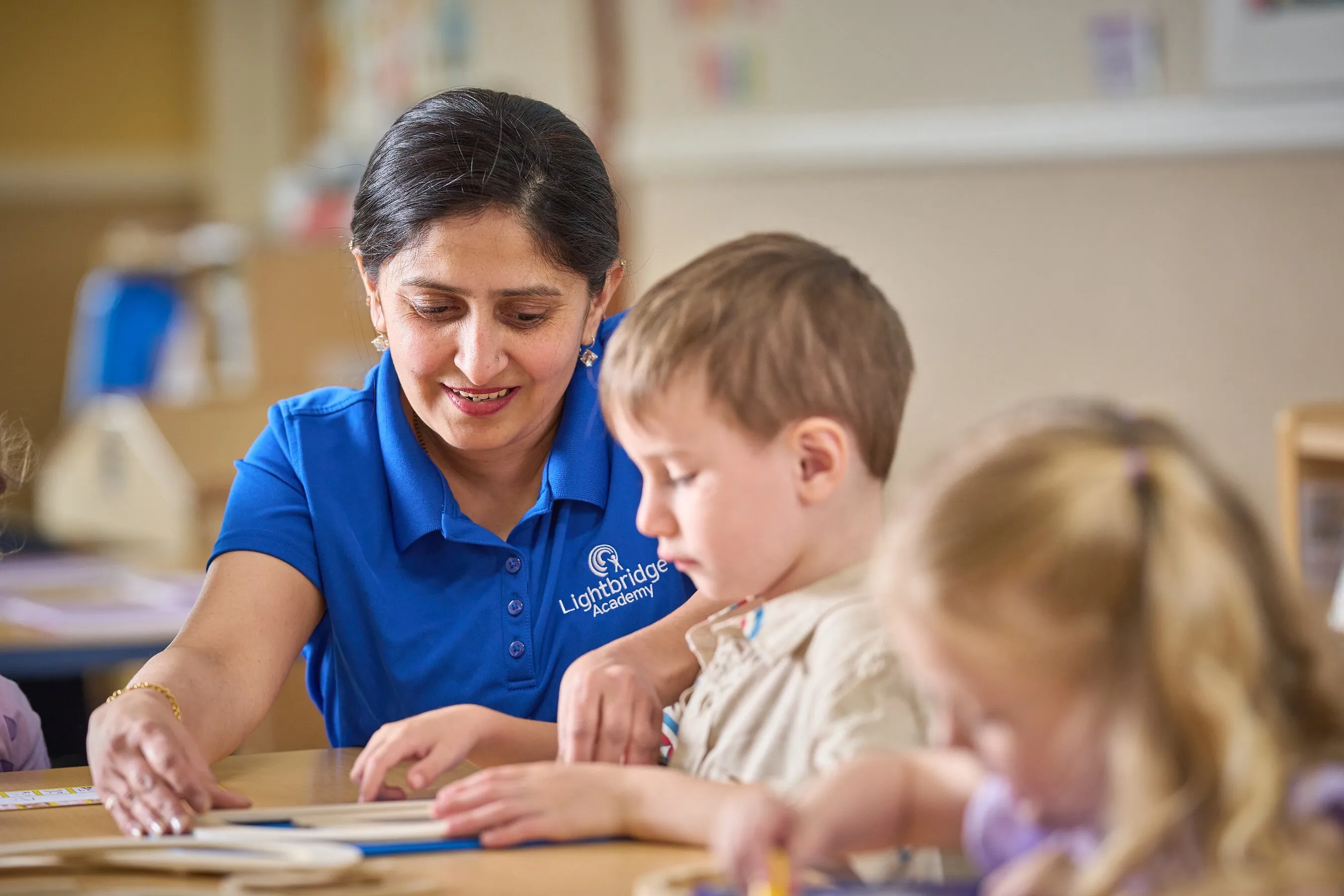 A teacher in a blue polo helps young children with an activity at a table.