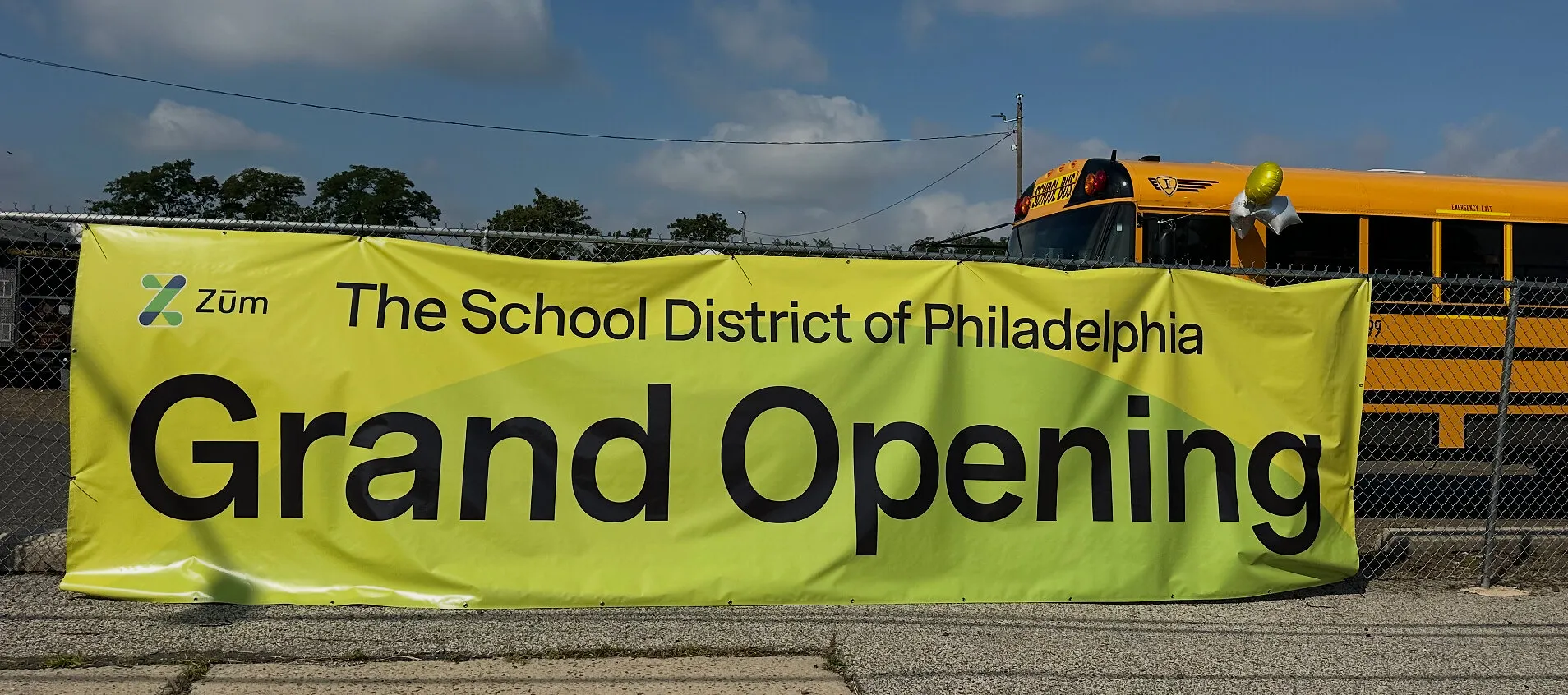 A Zūm and School District of Philadelphia grand opening banner hangs on a fence beside a school bus.