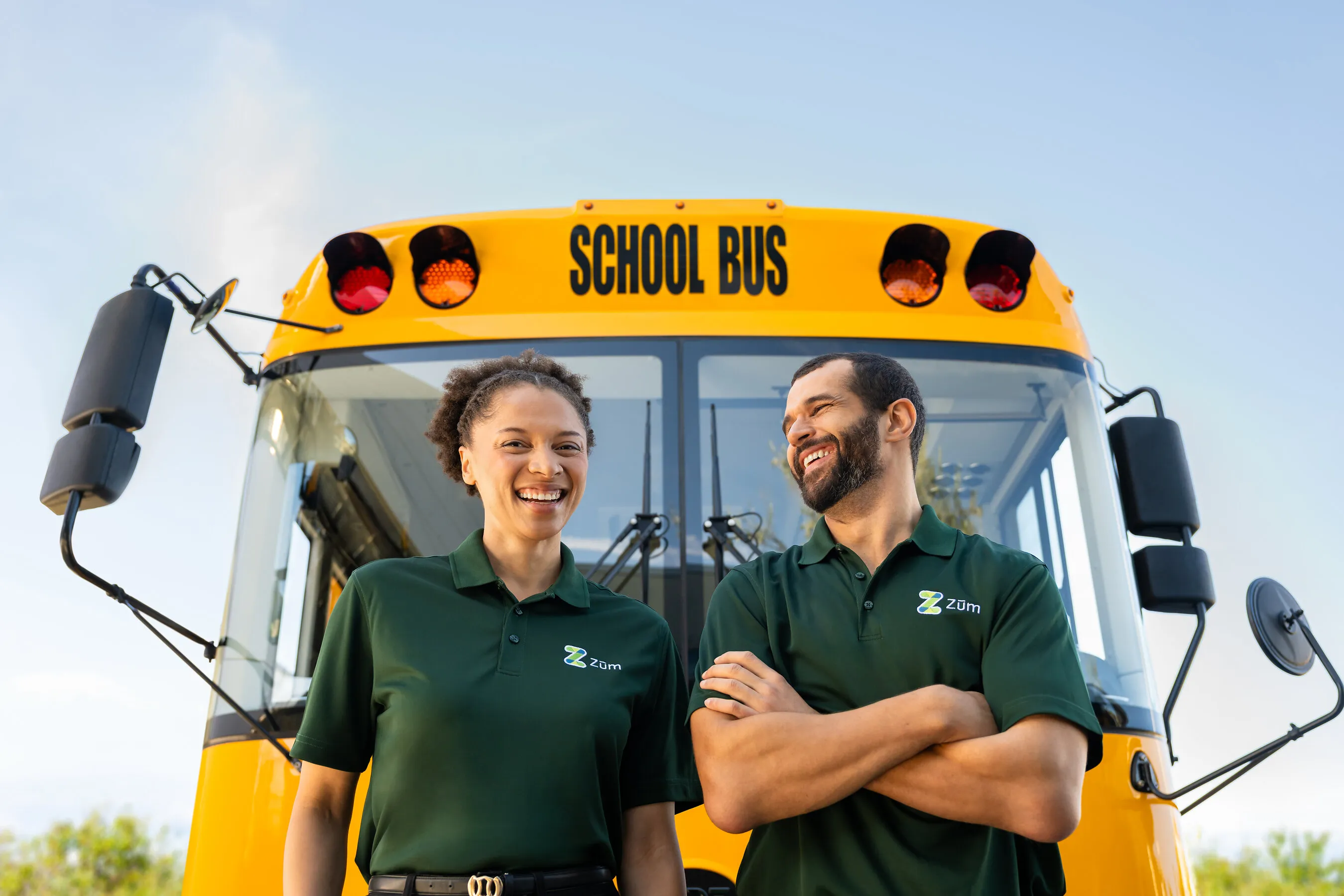 Two school bus drivers stand smiling in front of a yellow school bus.