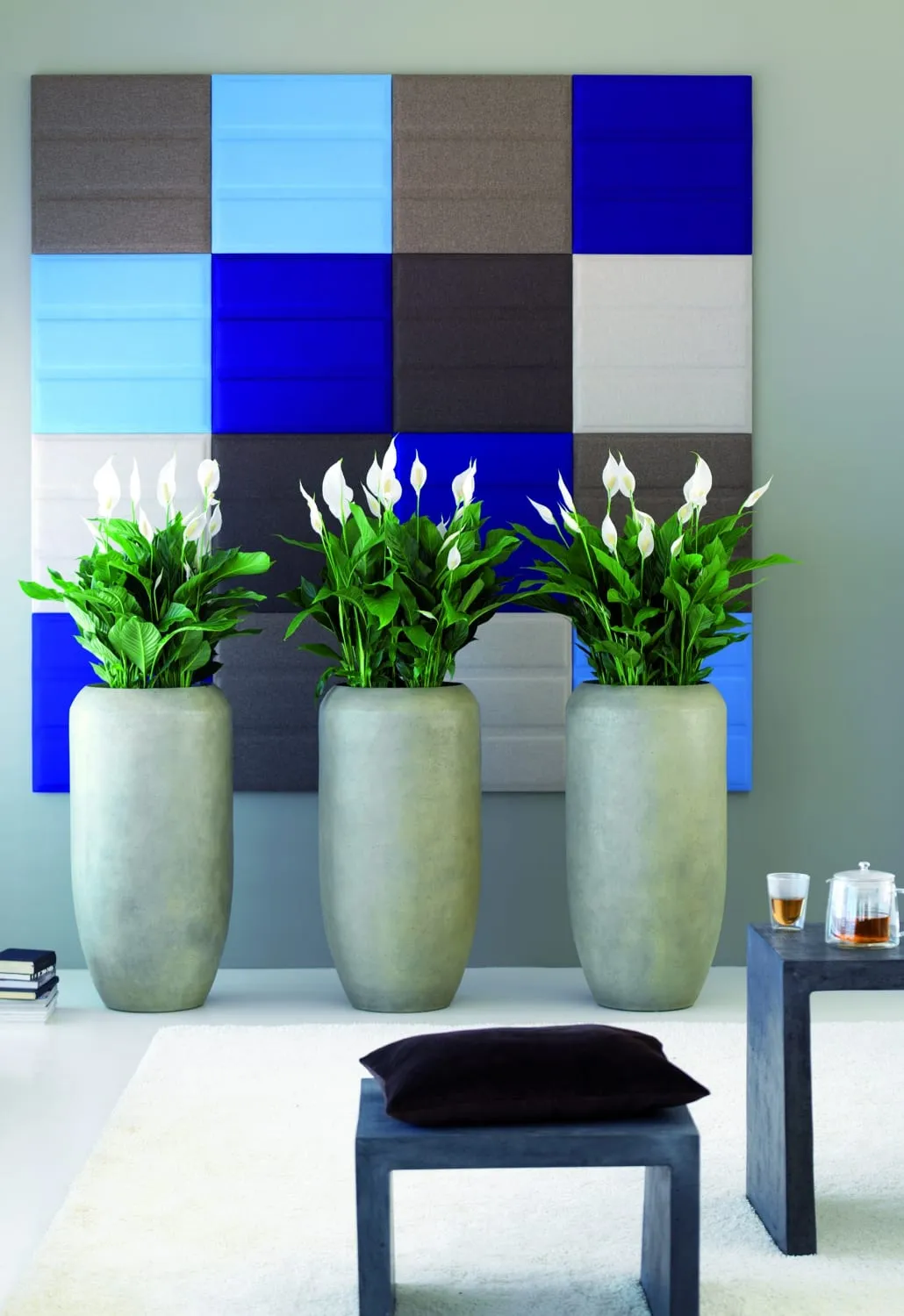 Three tall grey planters of white-flowered peace lilies stand in front of a wall of blue and grey fabric acoustic panels.