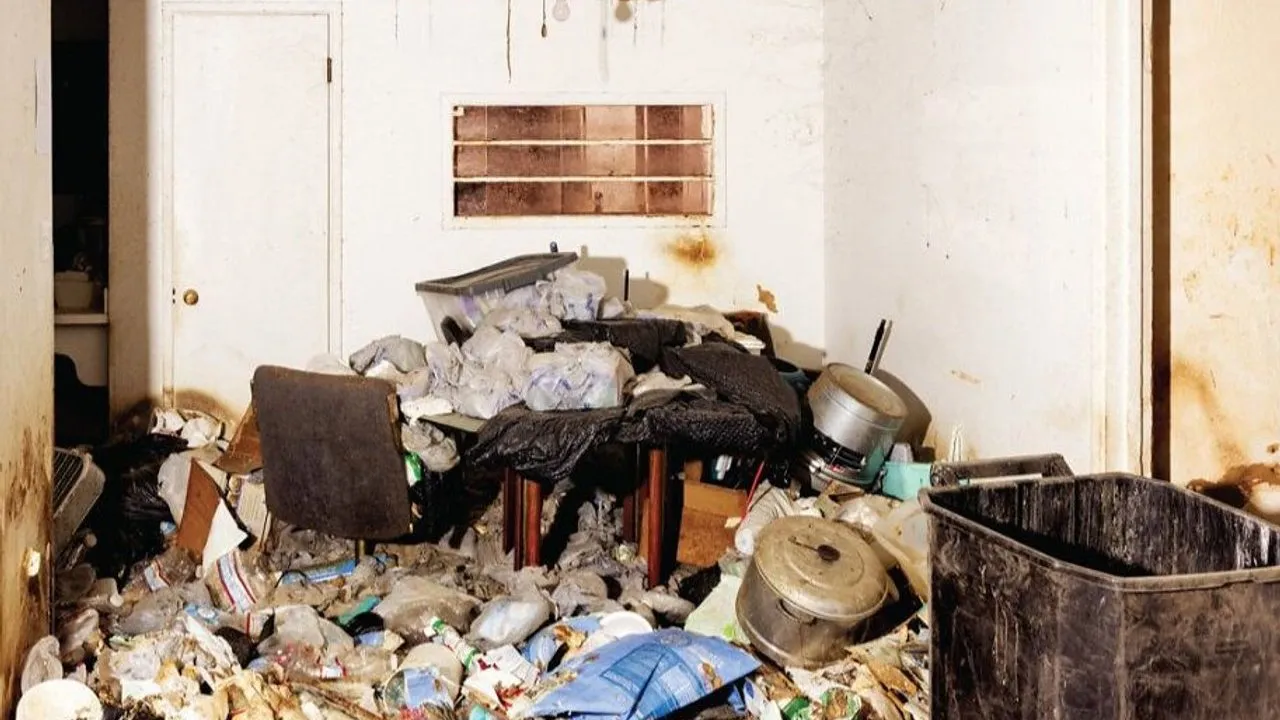 A cluttered room filled with trash, plastic bags, discarded furniture, pots and a large black bin.