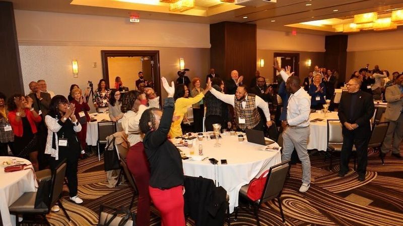 Conference attendees clap and high-five around tables in a hotel ballroom.