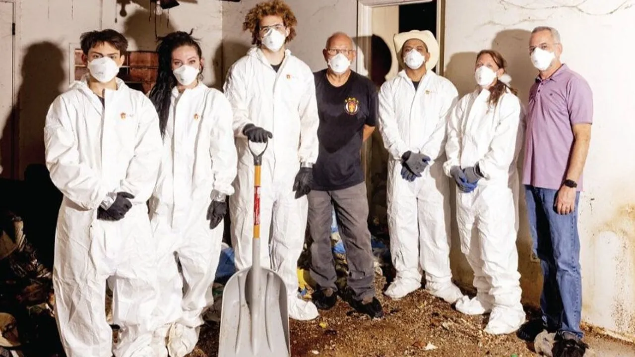 Seven people in masks, five in white protective suits, stand in a damaged room with a shovel.