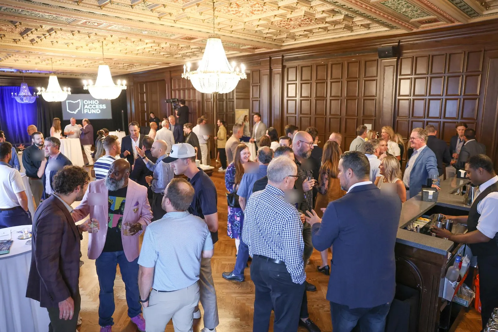 Guests stand and talk in a large wood-panelled ballroom with chandeliers and an ornate ceiling, with an Ohio Access Fund screen at the far end.