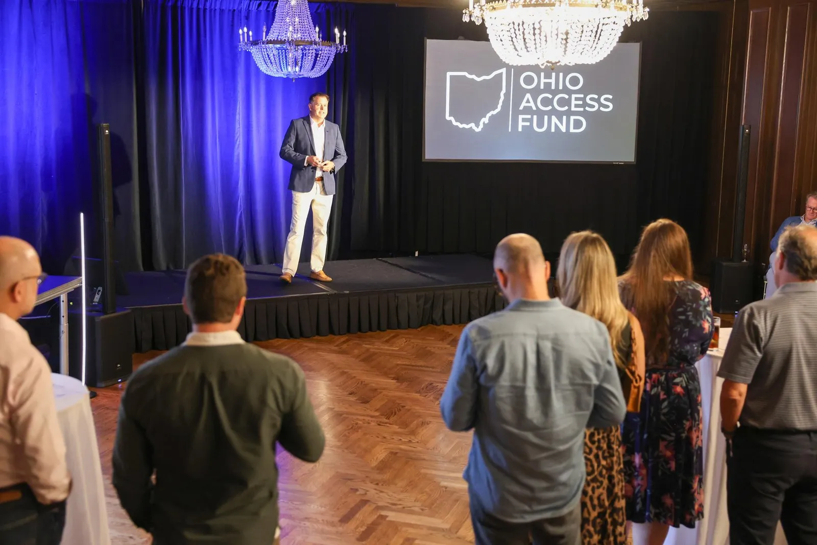 A speaker stands on a stage lit in blue beside an Ohio Access Fund logo screen as guests watch in a wood-floored ballroom.