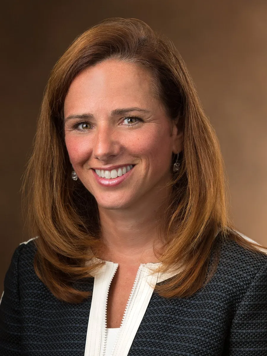 A smiling woman with brown hair wearing a black textured jacket with white trim.