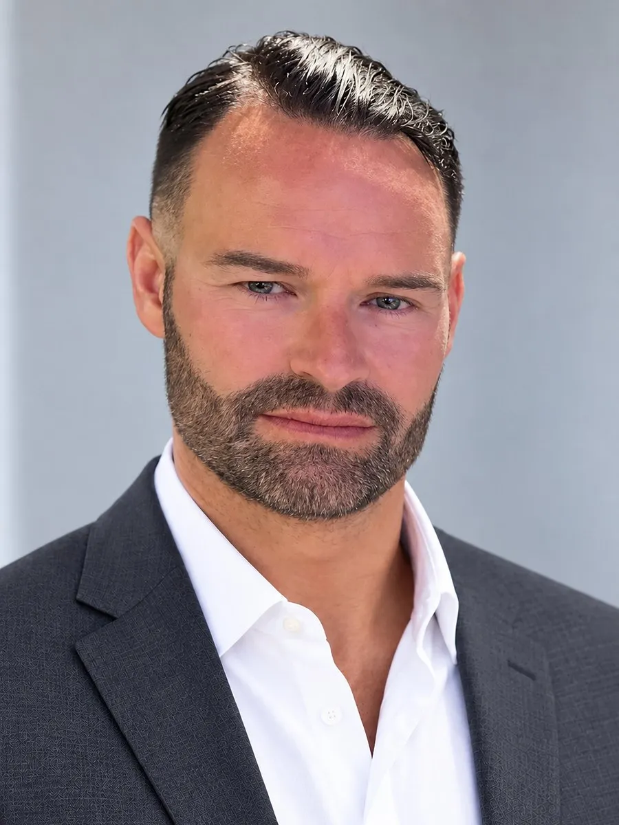 A bearded man in a dark suit jacket and white shirt against a gray background.