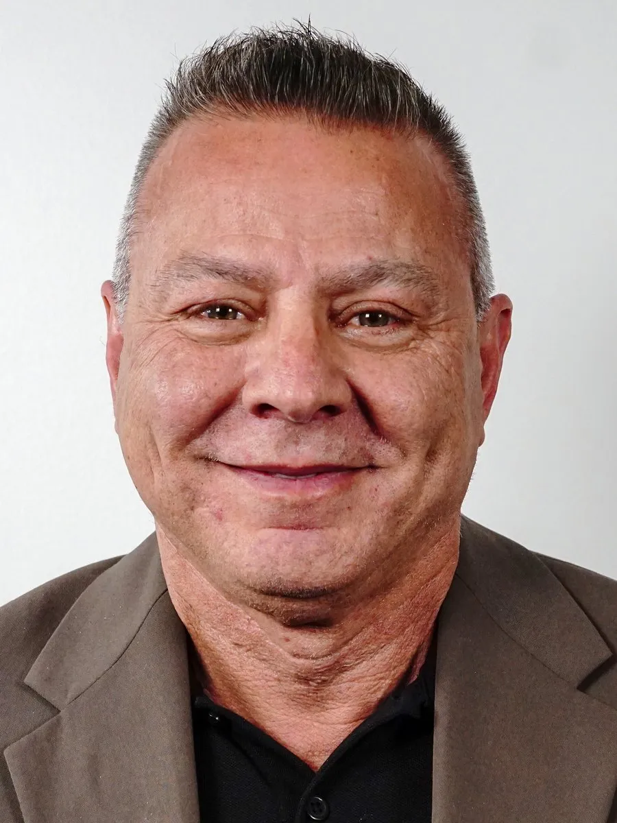 A close-up headshot of a smiling man with short grey hair in a brown jacket and black shirt.