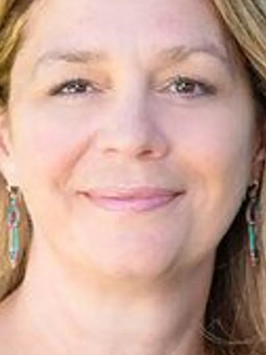A smiling blonde person wearing colorful dangling earrings outdoors.