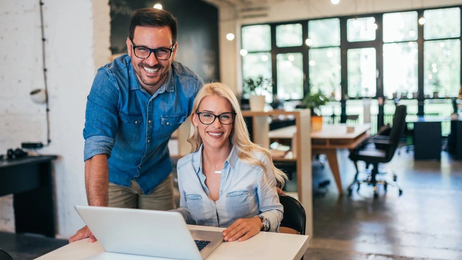 Two small business owners, a man and a woman, smile at the camera beside a laptop in their office.